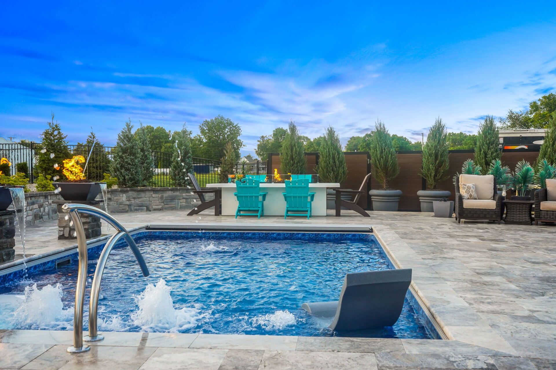 Outdoor hot tub with bubbling water, metal handrail, and lounge chairs on a sunny patio with greenery and blue sky