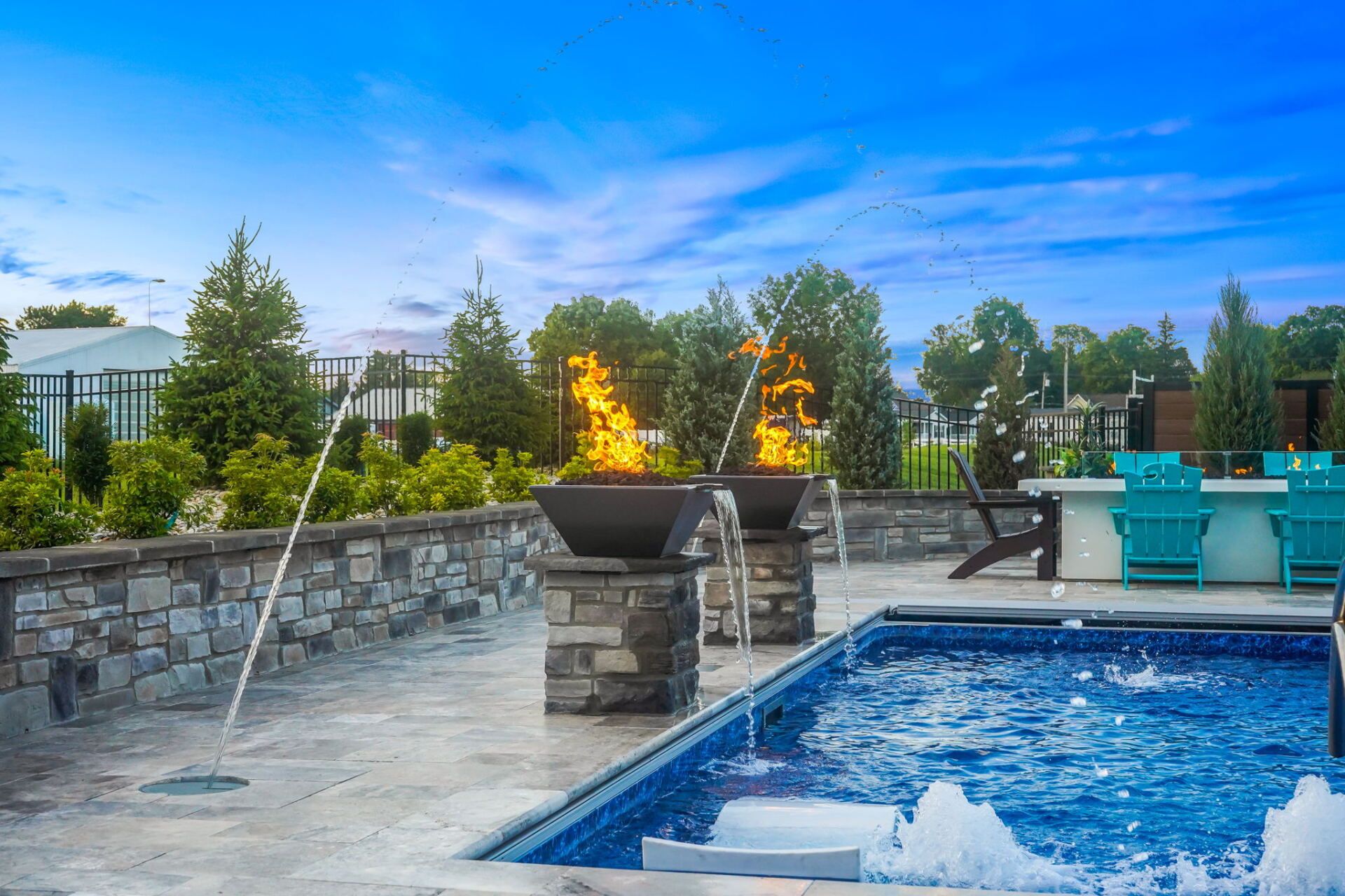 Backyard pool with stone patio, fire features, lounge chairs, and blue sky