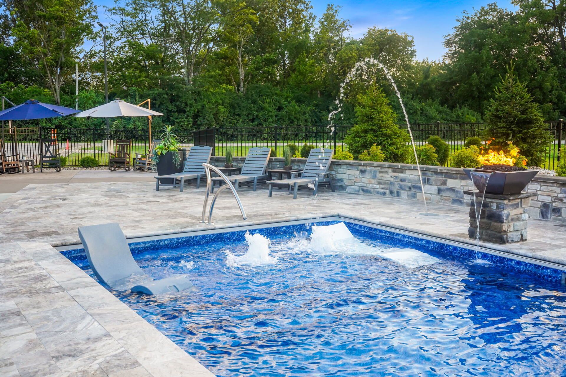 Backyard pool with blue water, lounge chairs, umbrellas, and a stone fire feature at dusk