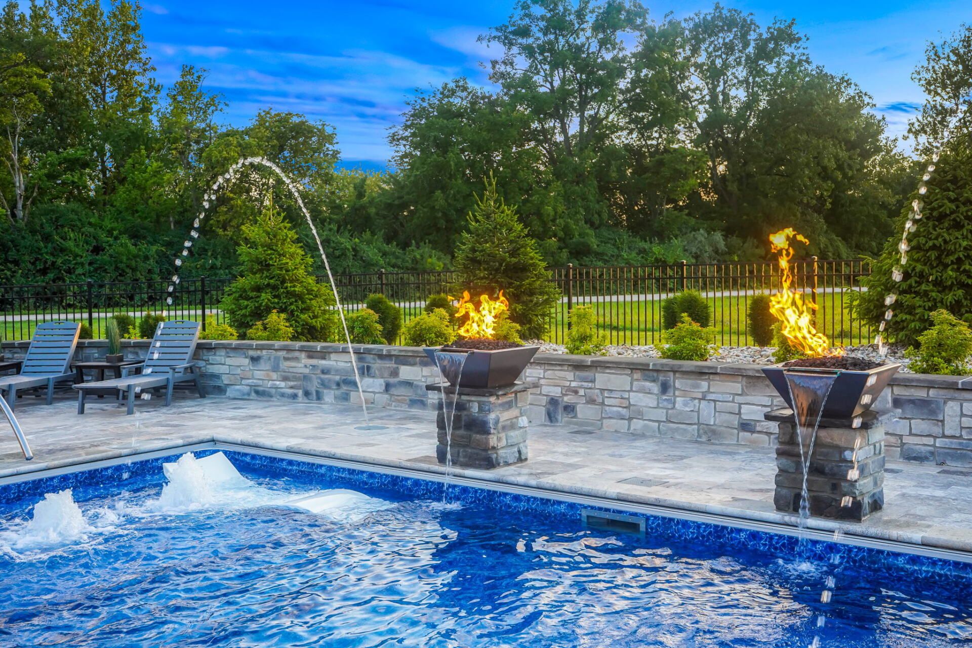Backyard pool with blue water, stone deck, lounge chairs, and lit fountains at dusk
