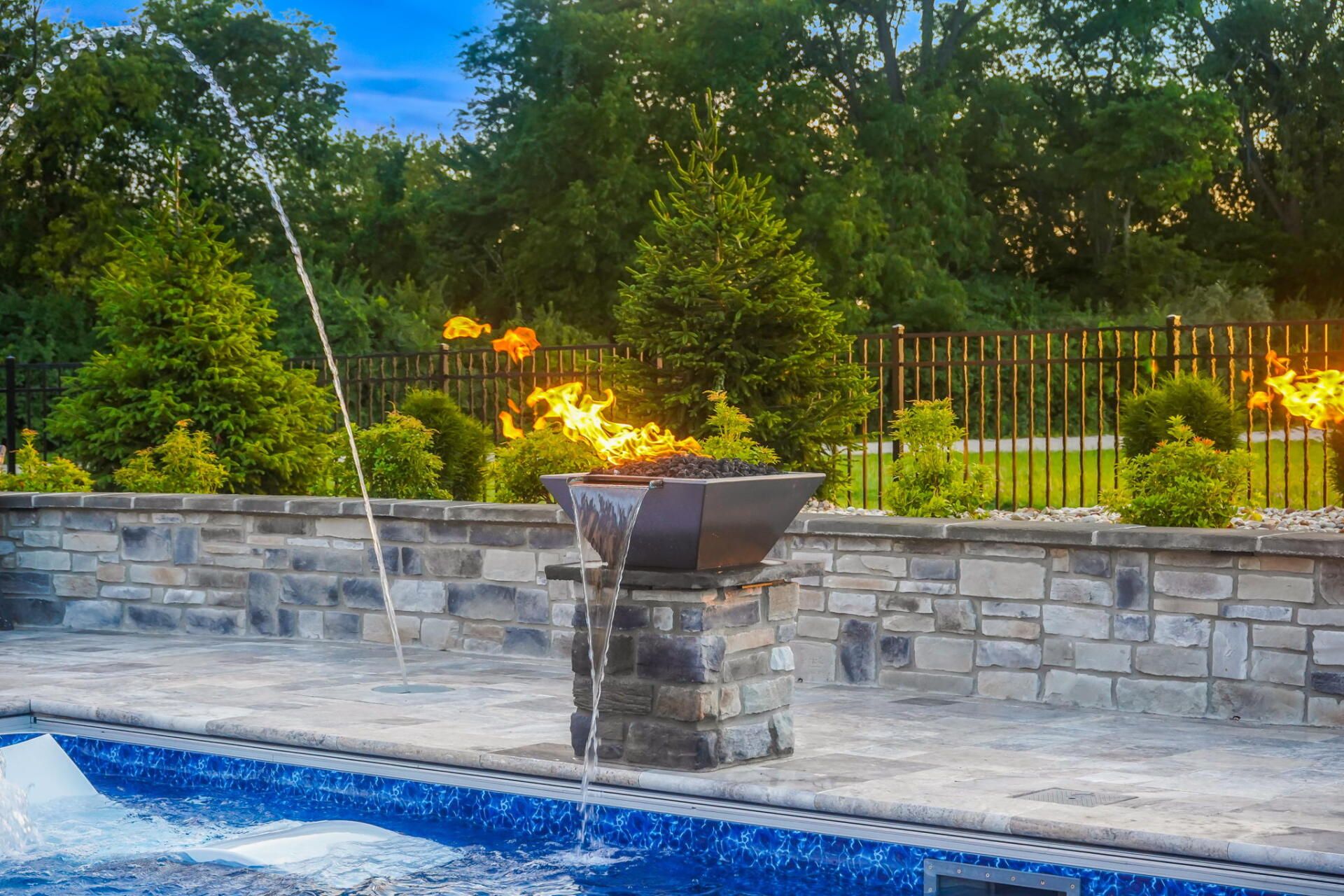 Backyard pool with a lit stone fire bowl, stone wall, and lush green trees at dusk