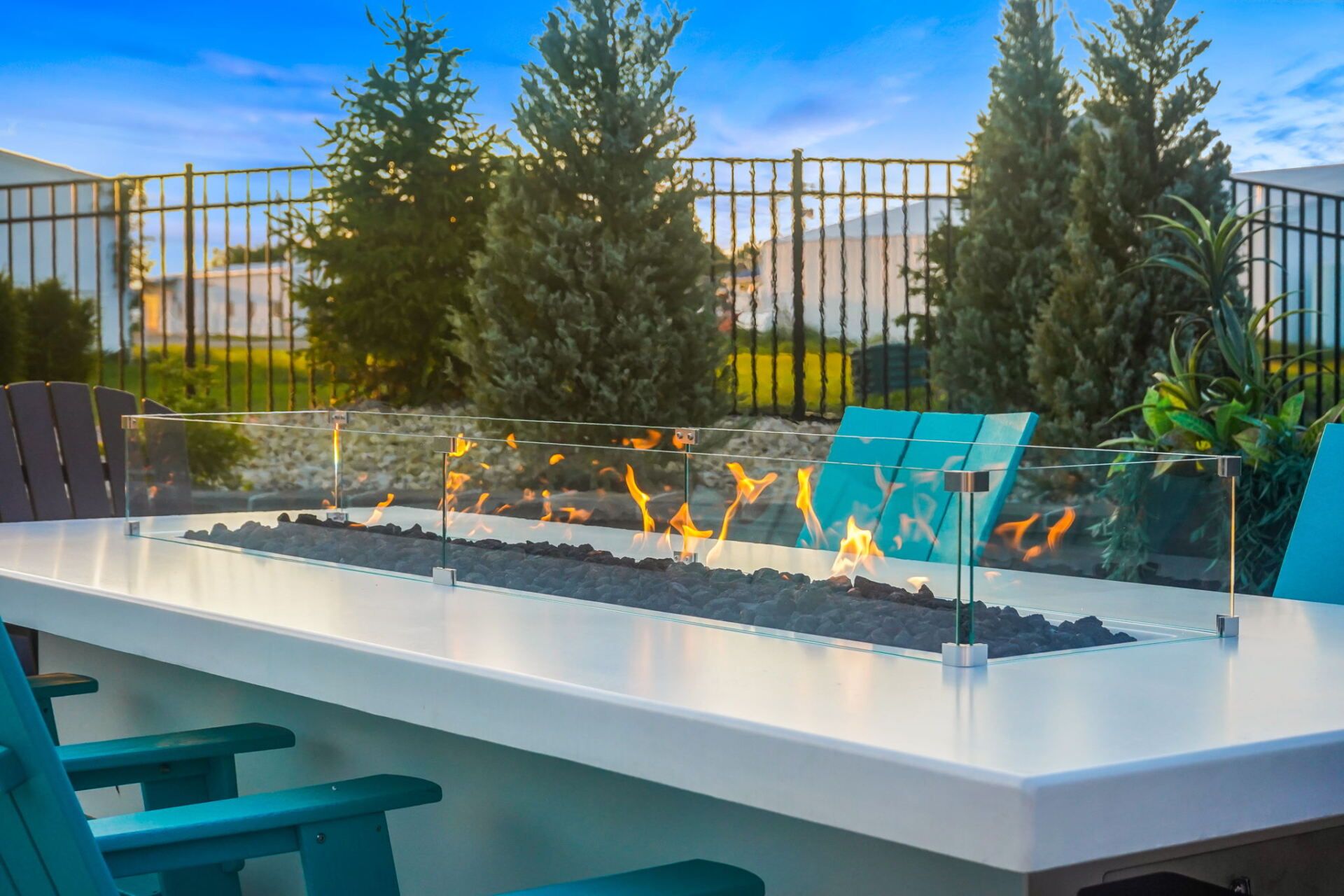 Modern patio with a rectangular fire pit, turquoise chairs, and a fenced garden at sunset