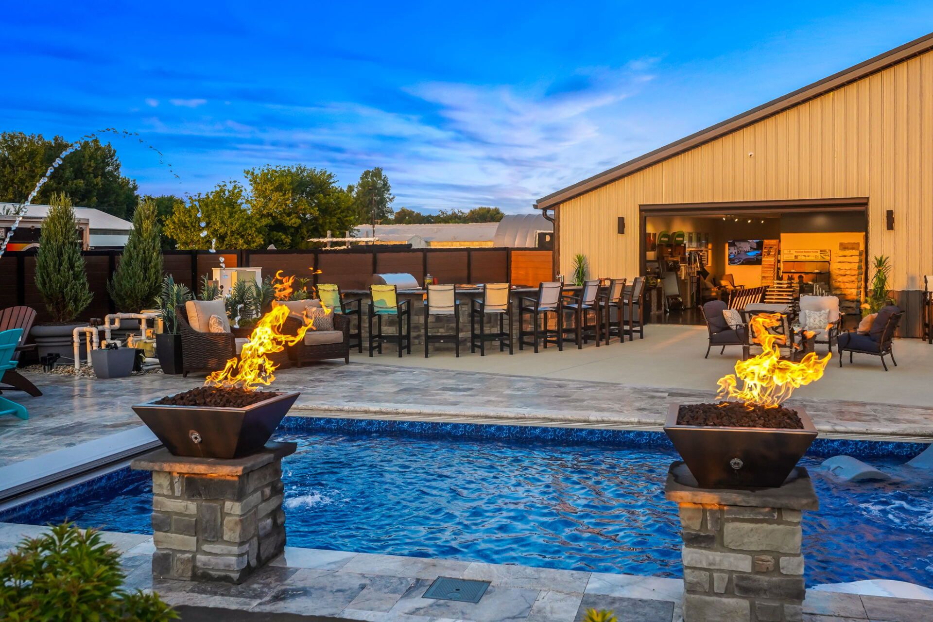 Outdoor pool with fire pits and dining area beside a tan building at dusk