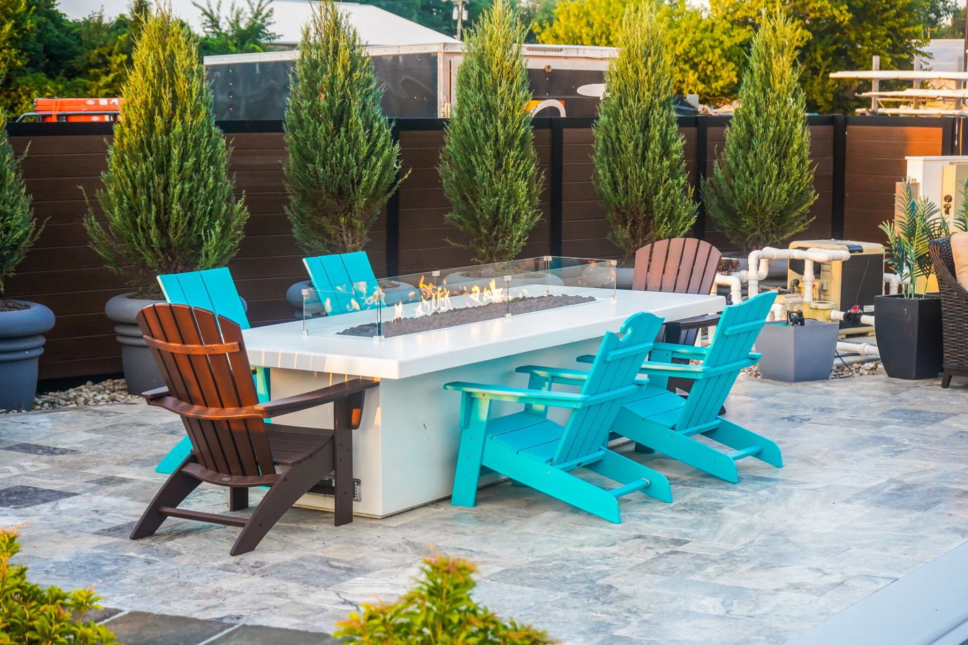 Outdoor patio with a fire pit table, turquoise chairs, and tall potted evergreens under warm sunlight