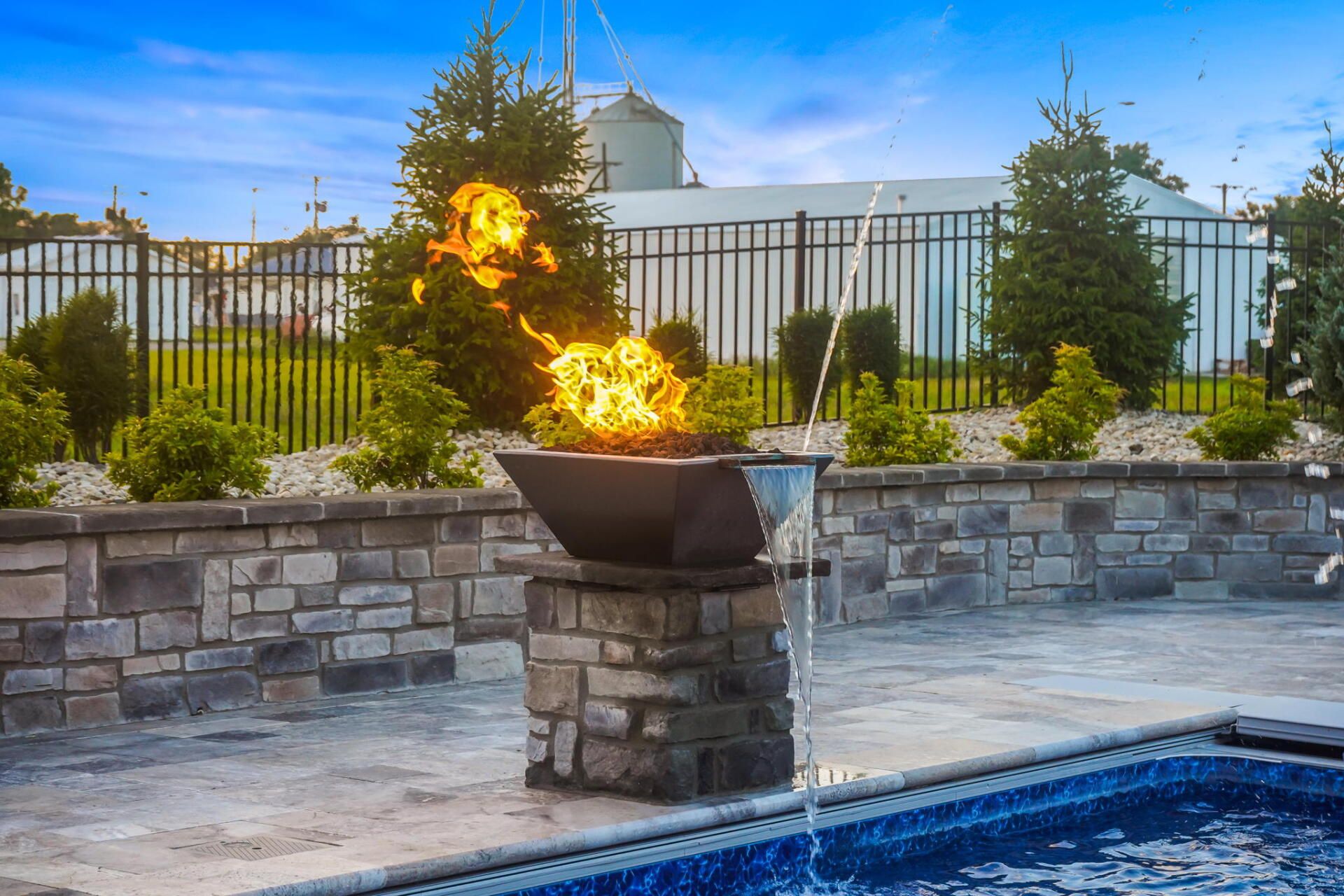 Backyard poolside fire pit with flames, stone wall, fence, and manicured shrubs