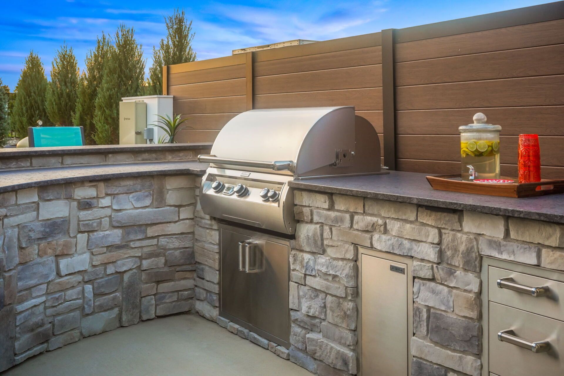 Outdoor stone patio kitchen with built-in grill, counter, cabinets, and a wooden privacy fence