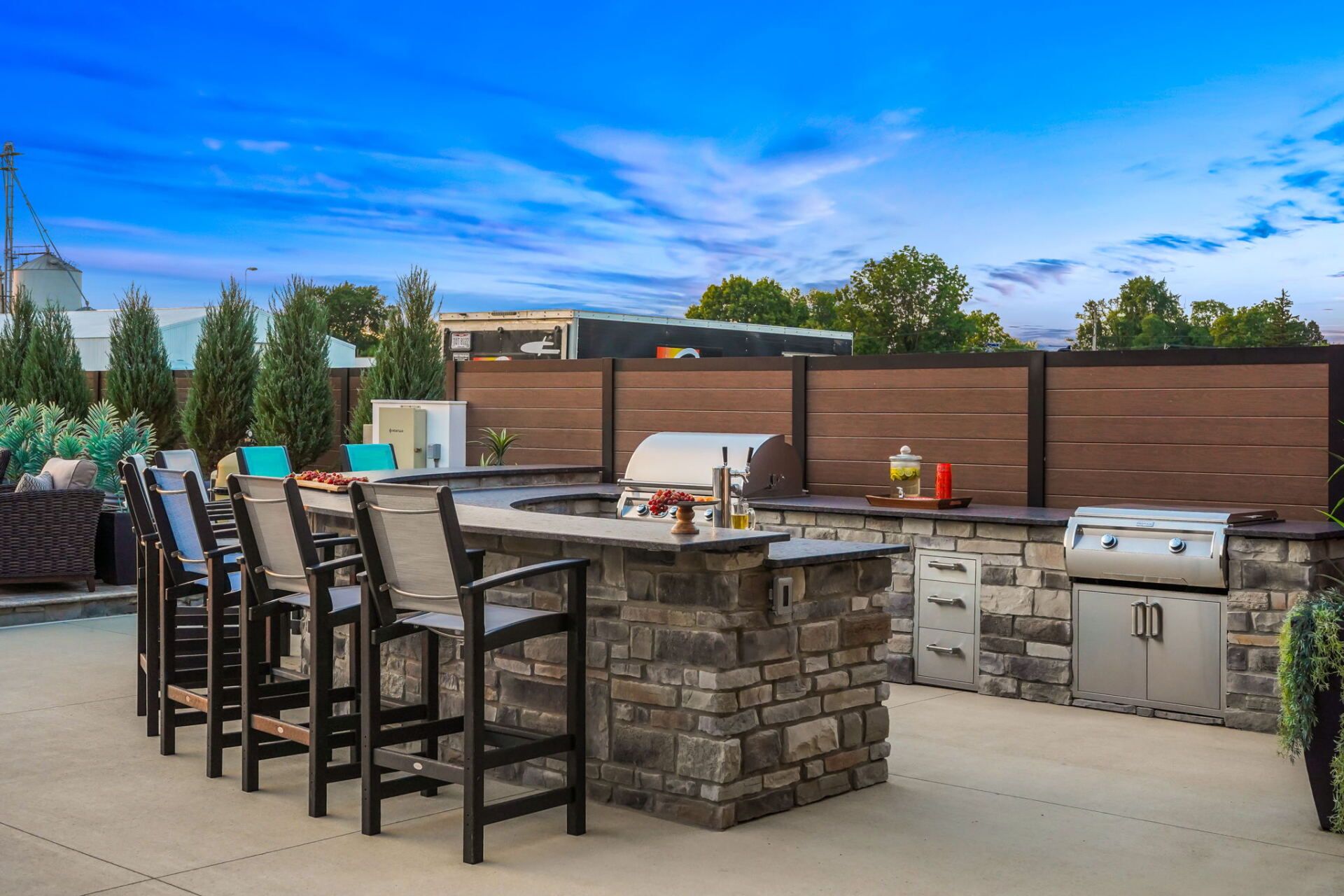 Backyard outdoor kitchen with stone island, grill, bar stools, and wooden privacy fence under a blue sky