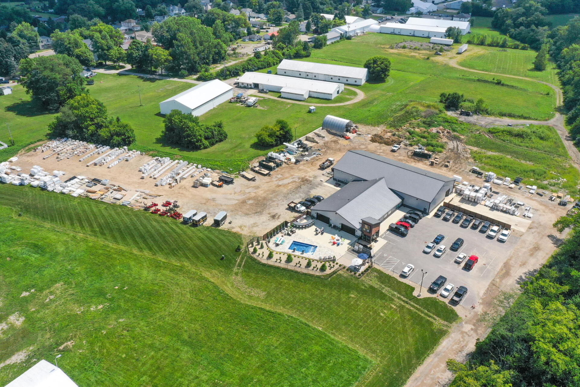 Aerial view of a rural construction site with buildings, parked vehicles, and green fields nearby