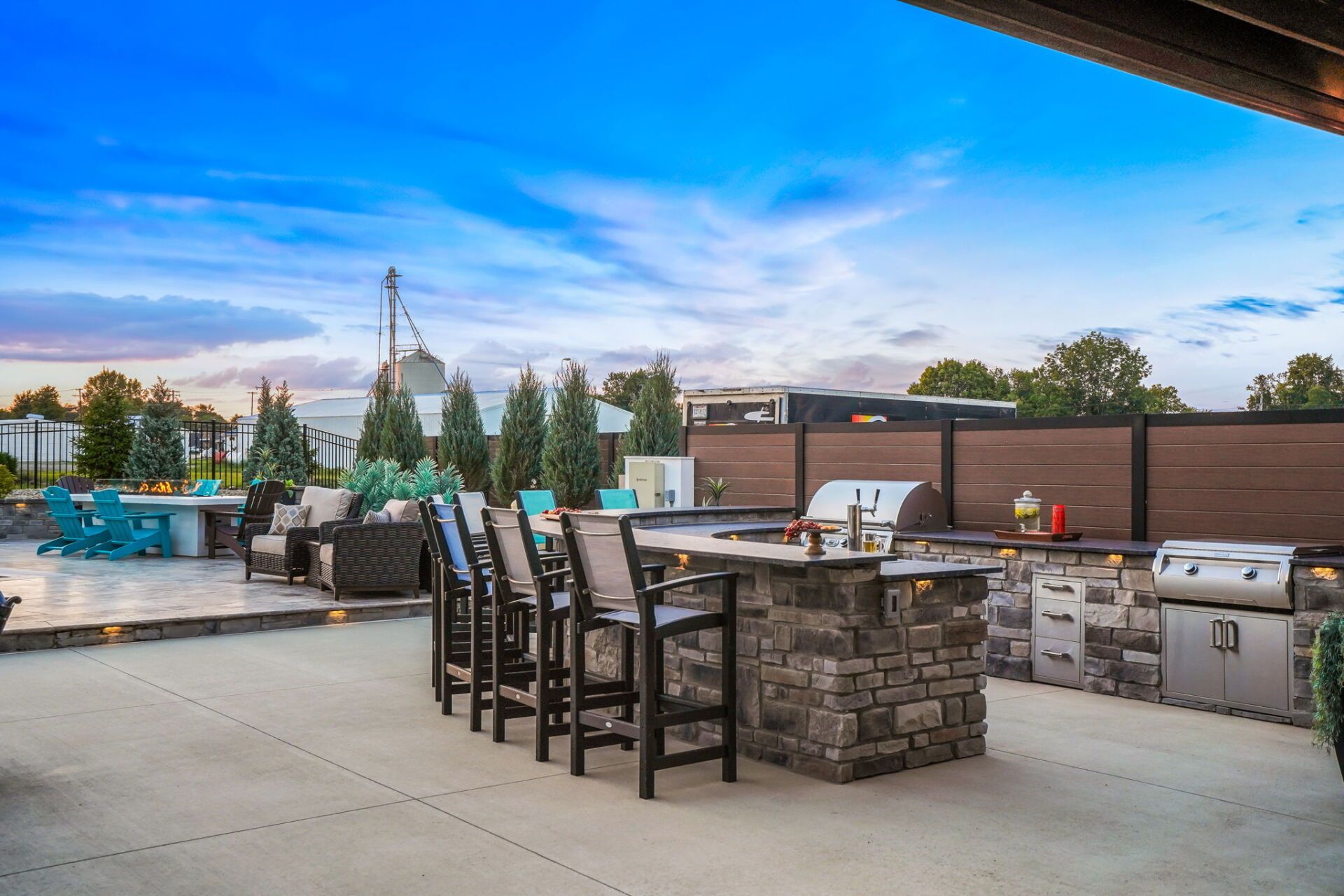 Outdoor patio with bar stools, stone fire pit, and grill under a blue sky