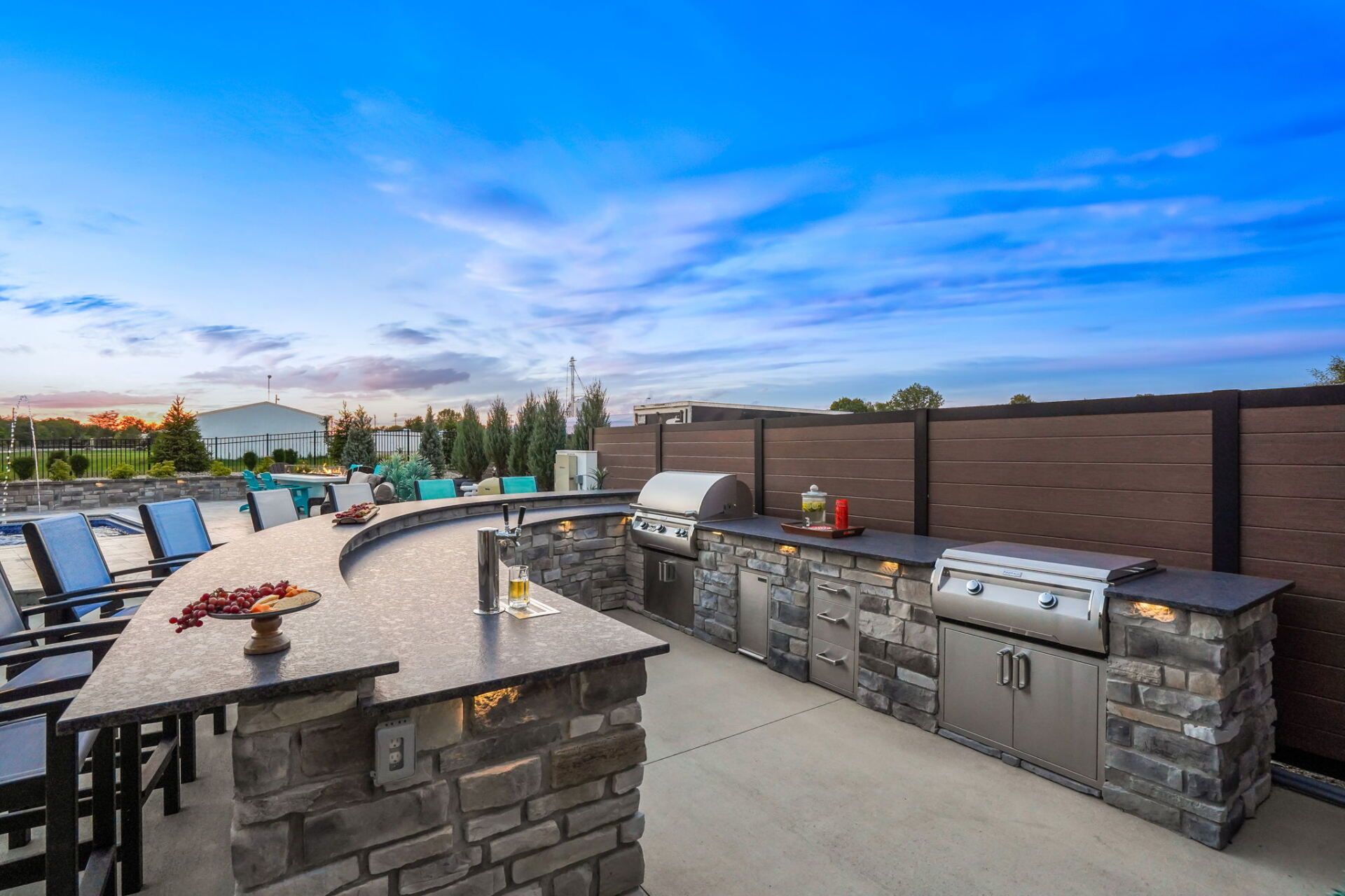 Outdoor stone patio with built-in grill and fire pit, lined with lounge chairs under a blue sky