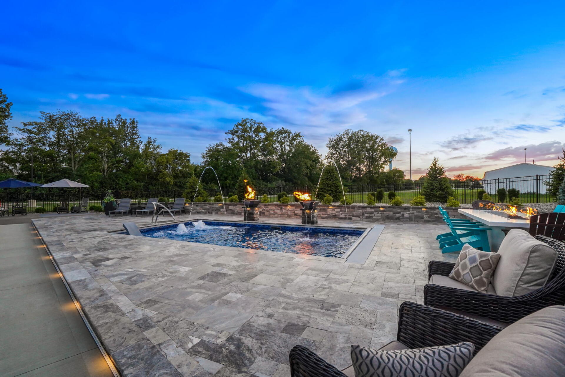 Stone patio with a central fire pit, wicker seating, and a backyard pool at dusk