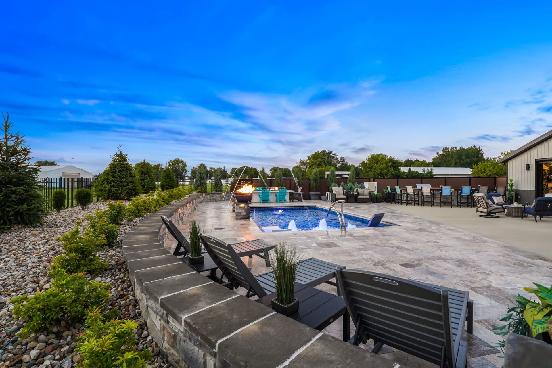 Backyard pool with blue water, stone patio, lounge chairs, and potted plants under a bright sky