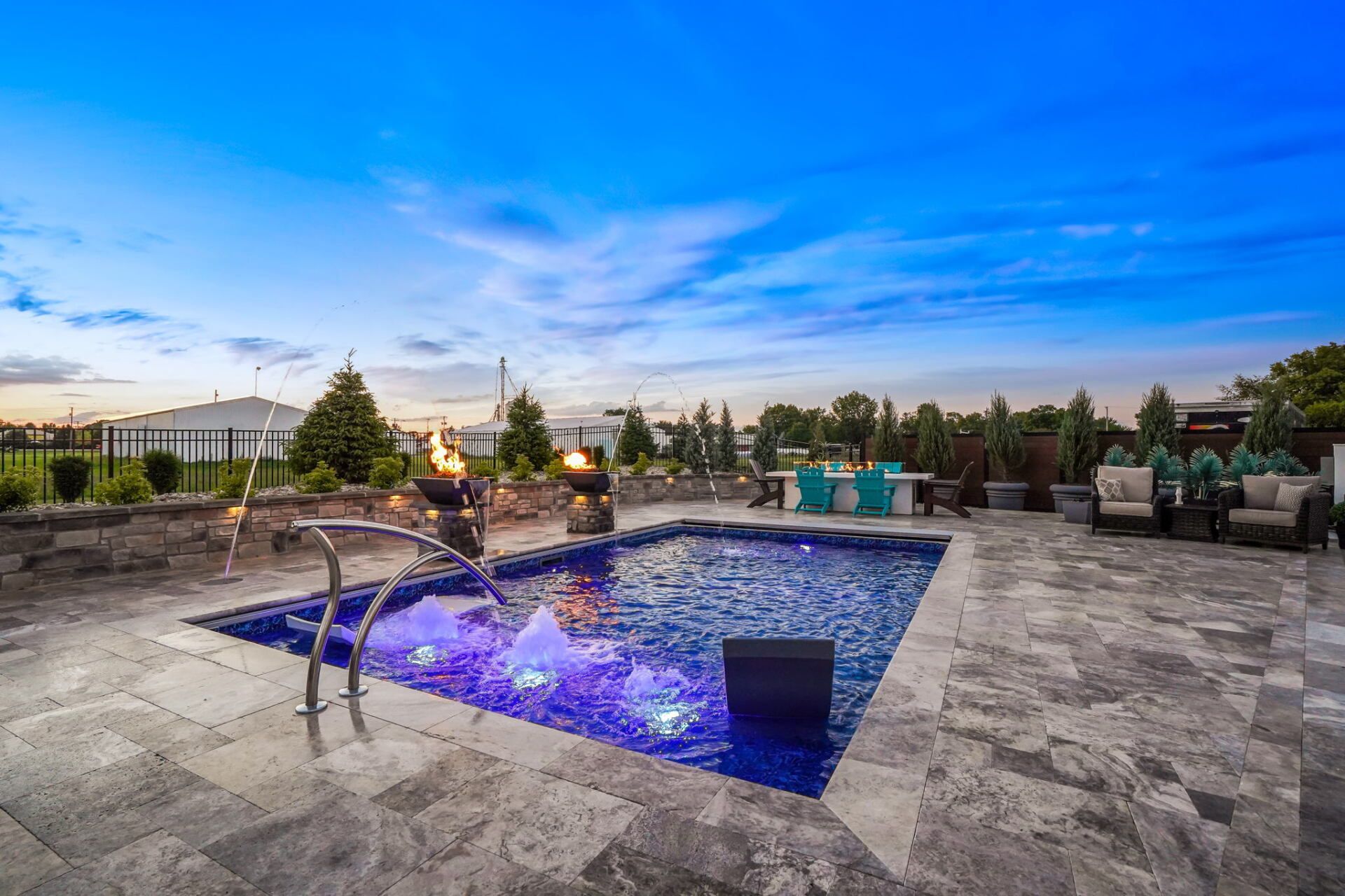 Modern backyard patio with a lit square pool, stone decking, and lounge seating at dusk