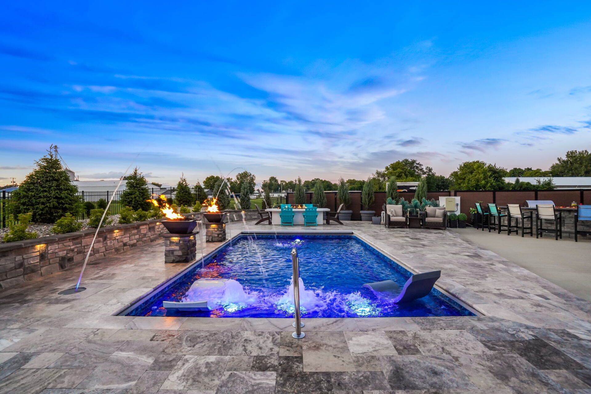 Lit backyard pool at dusk with stone patio, lounge chairs, and a blue sky