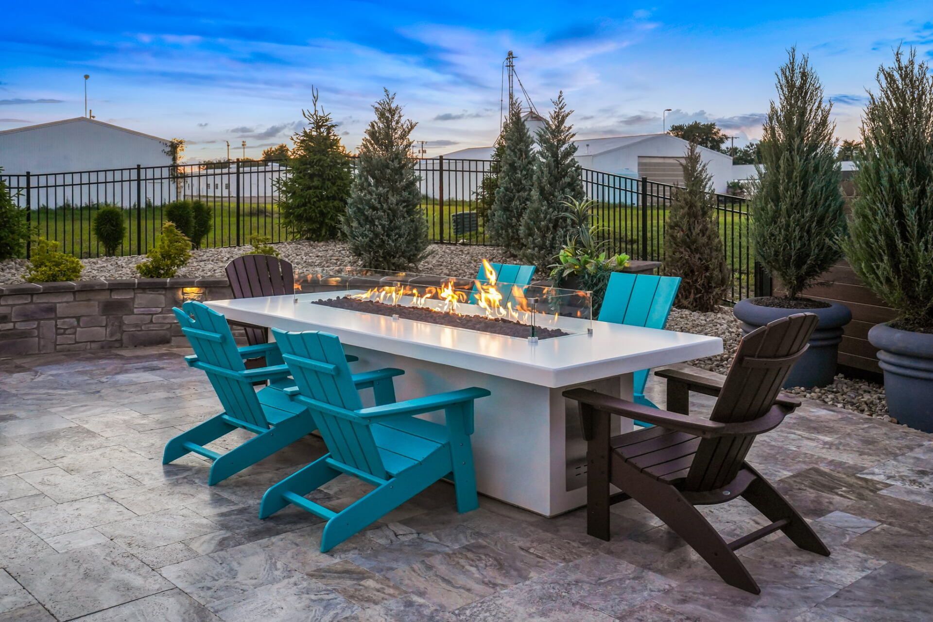 Outdoor patio with turquoise chairs around a fire table, surrounded by potted evergreens and stone landscaping.