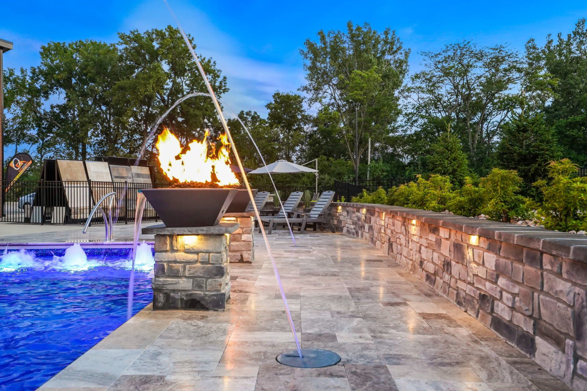 Backyard pool patio with fire feature, stone wall, and water jets at dusk