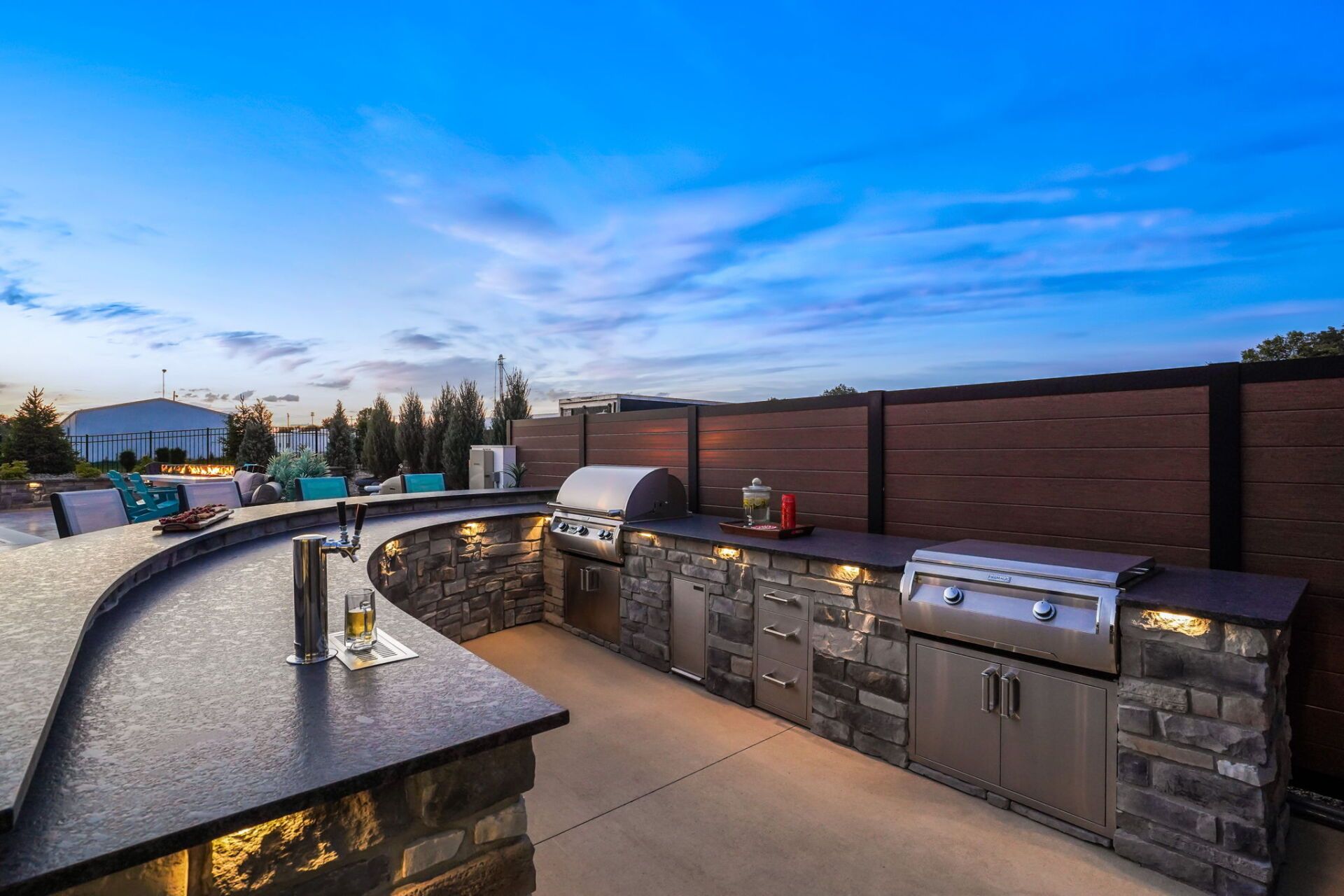 Outdoor stone patio with curved grill island, built-in sink, and blue evening sky
