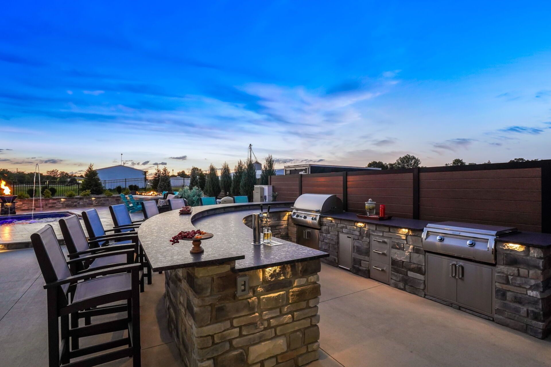Outdoor patio with stone bar, chairs, and built-in grill at dusk under a blue sky