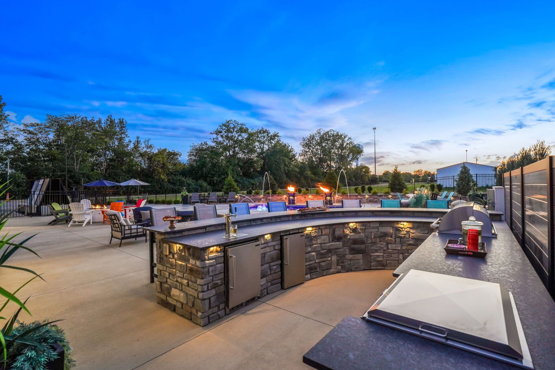 Outdoor patio bar at dusk with stone counter, red stools, and warm lights under a blue sky