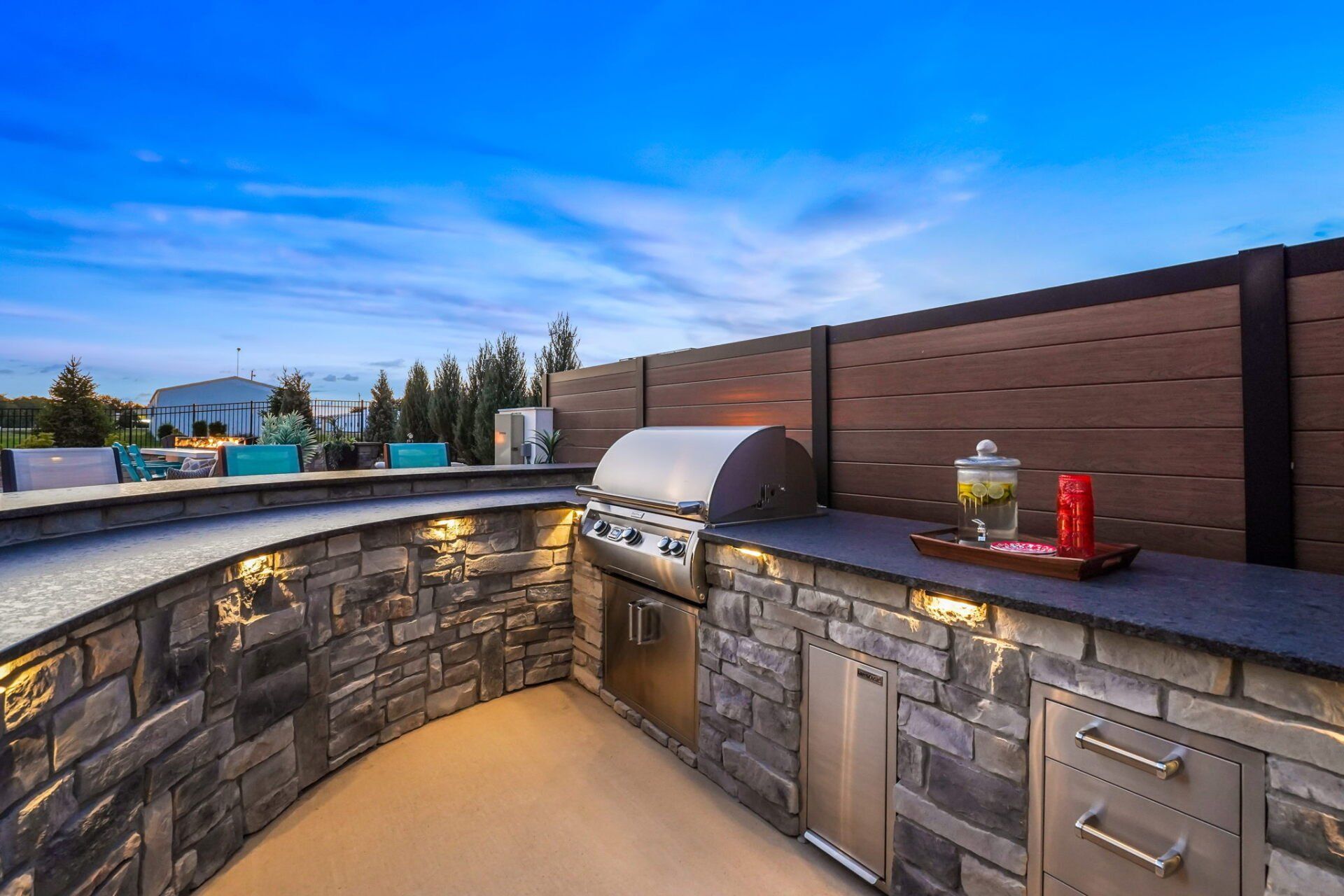 Outdoor stone barbecue counter with grill, sink, and red condiment bottles at dusk