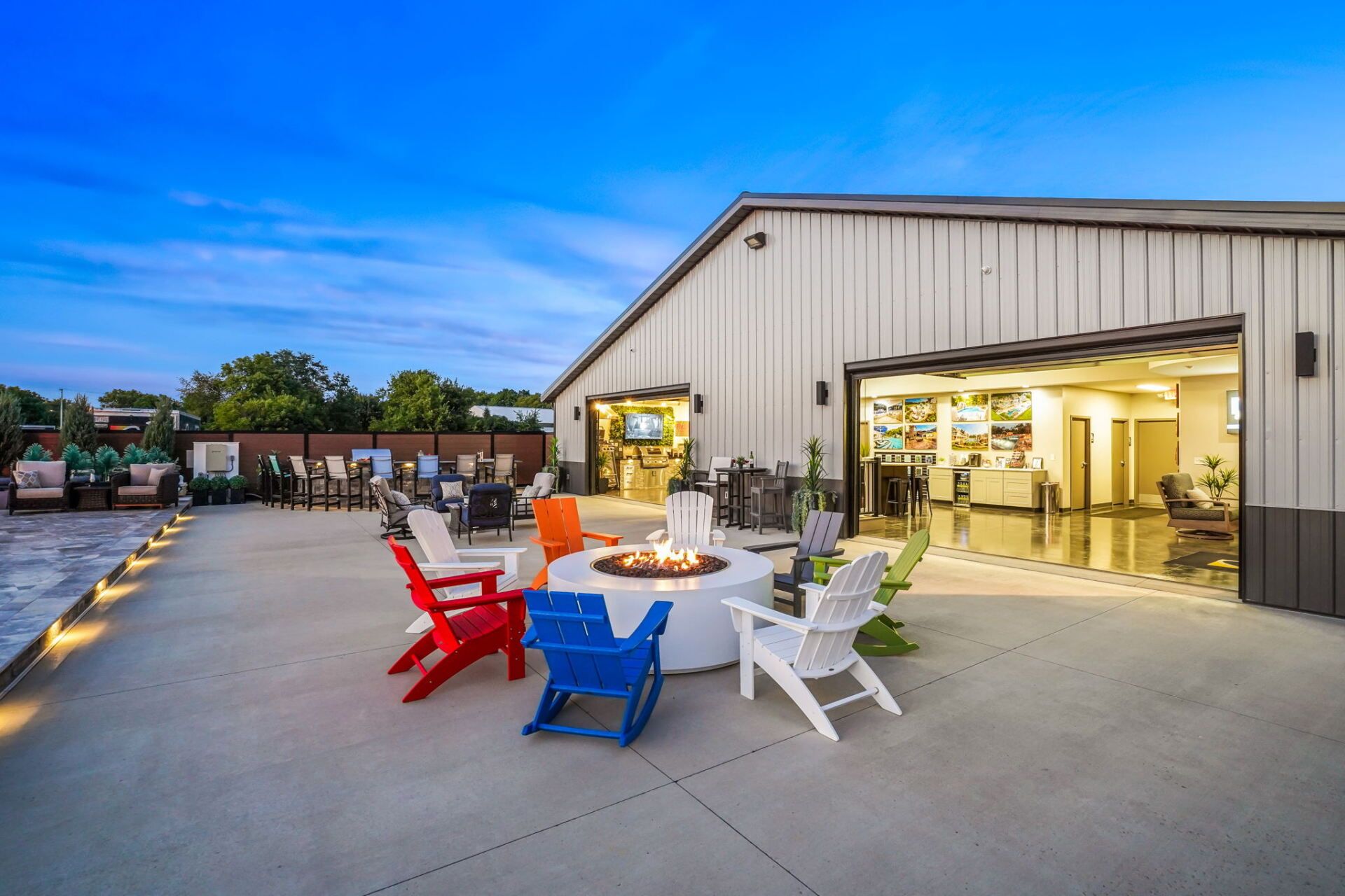Outdoor patio with colorful chairs and a fire pit beside an open industrial-style building at dusk