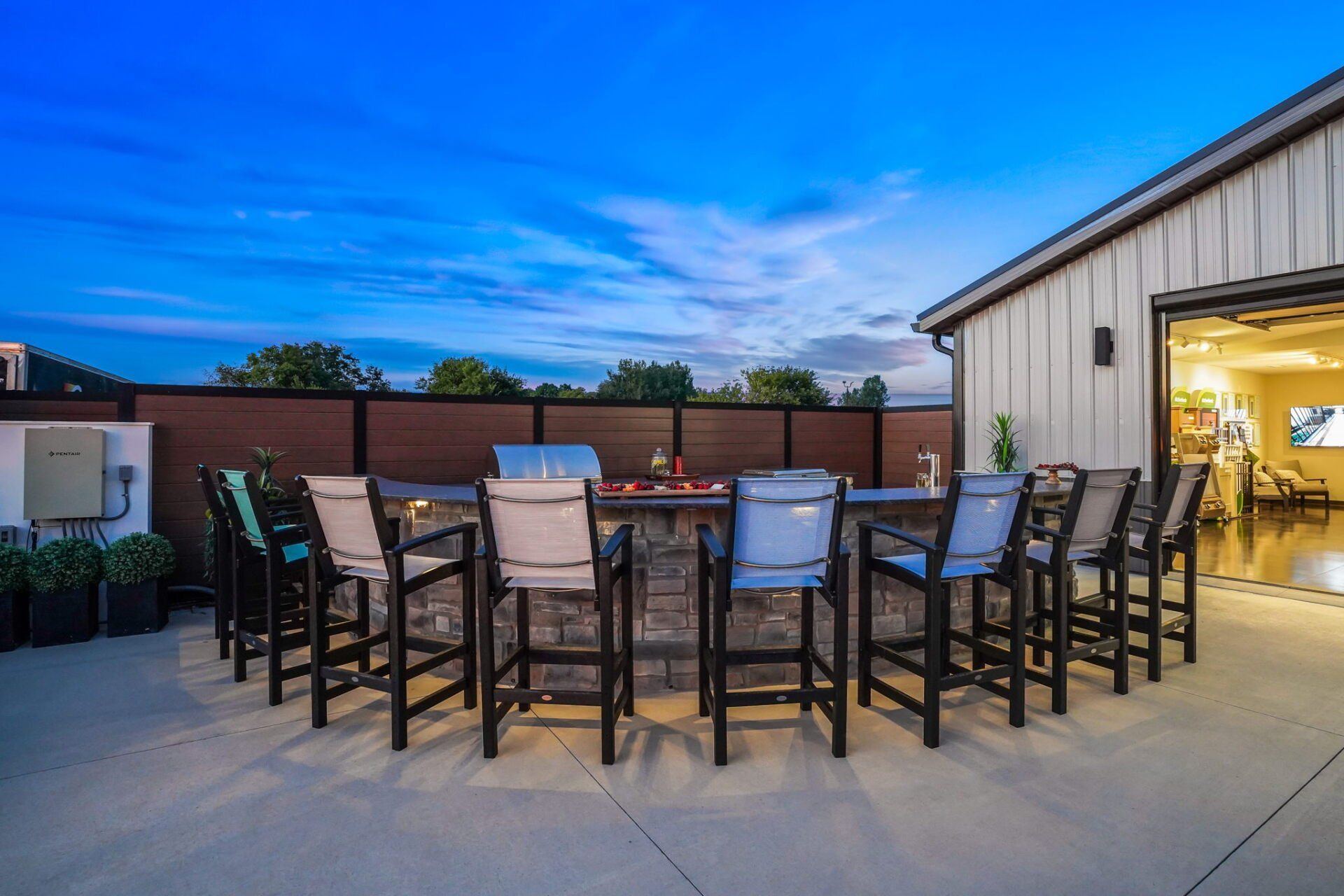 Outdoor patio with bar stools around a fire pit beside an open garage at dusk