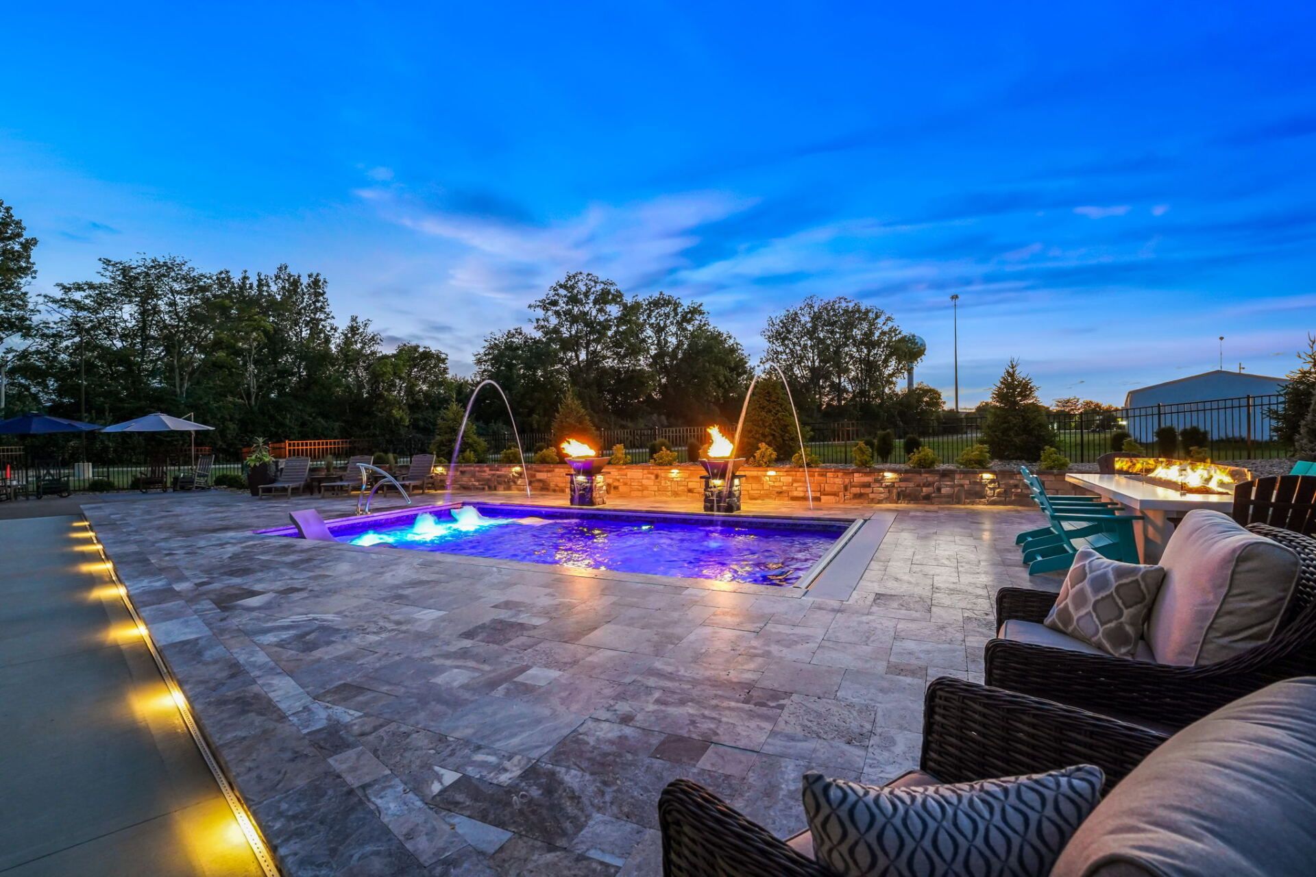 Outdoor pool patio at dusk with blue-lit water, fire torches, and lounge seating under a twilight sky
