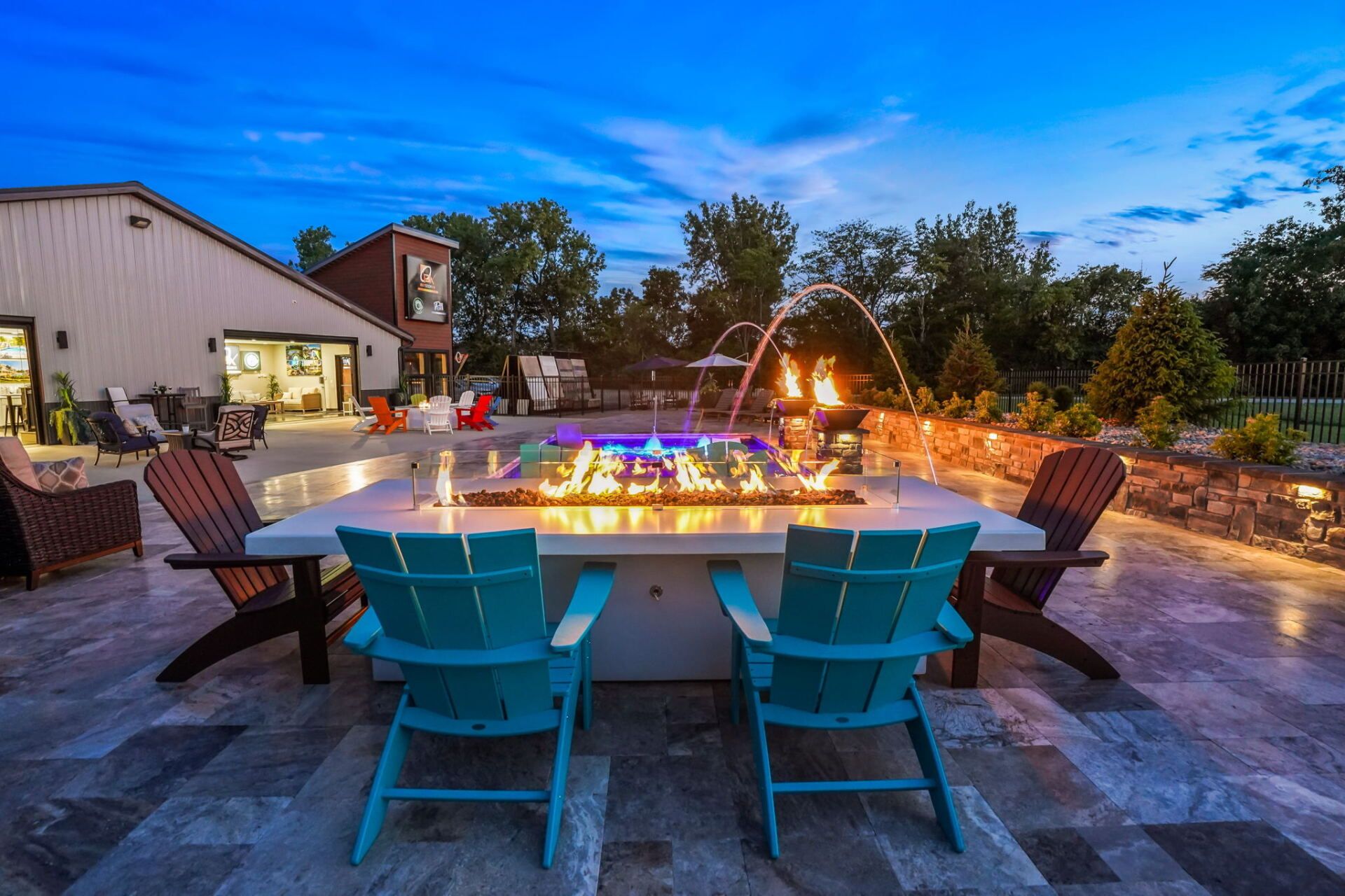 Backyard patio with turquoise chairs around a lit fire pit at dusk, beside a house and stone wall