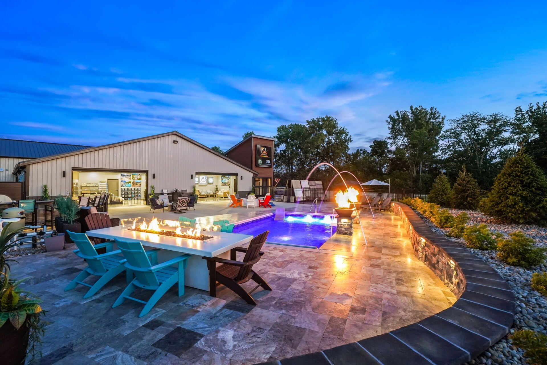 Backyard patio with lit pool, turquoise chairs, fire pit, and modern house at dusk