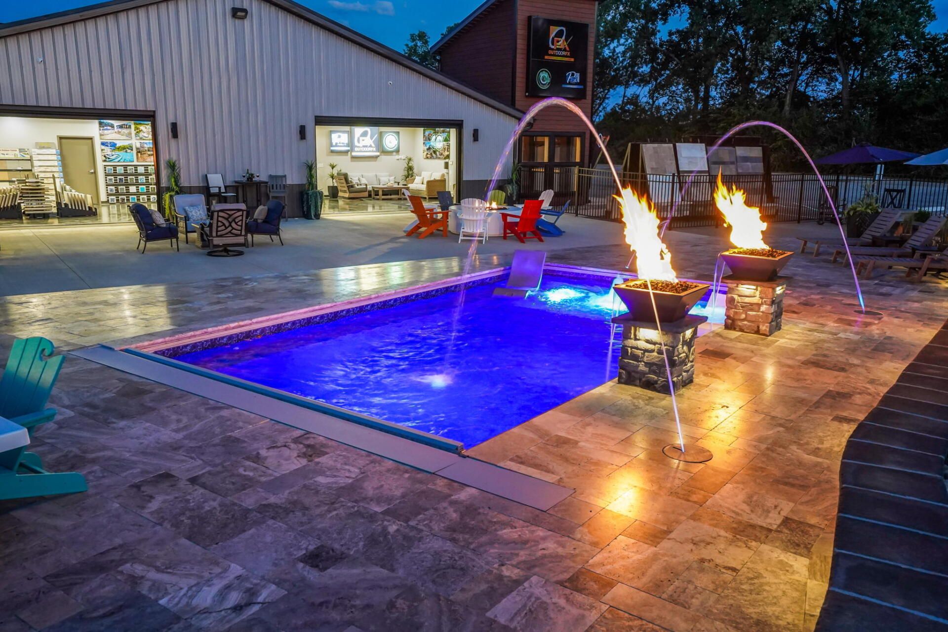 Backyard pool at night with blue-lit water, lounge chairs, and fire bowls on the patio