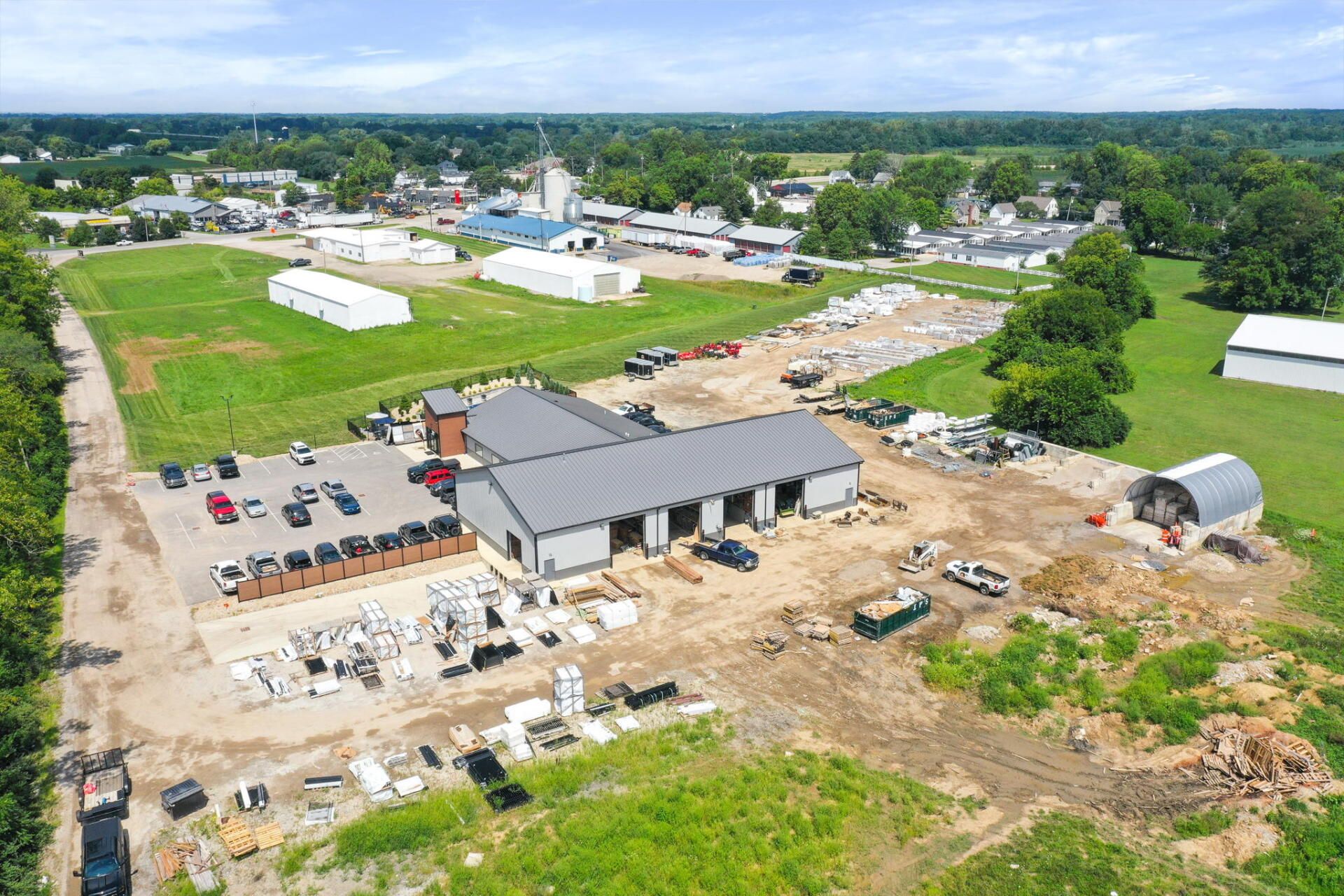Aerial view of a construction site with a gray building, dirt lot, parked cars, and green fields nearby