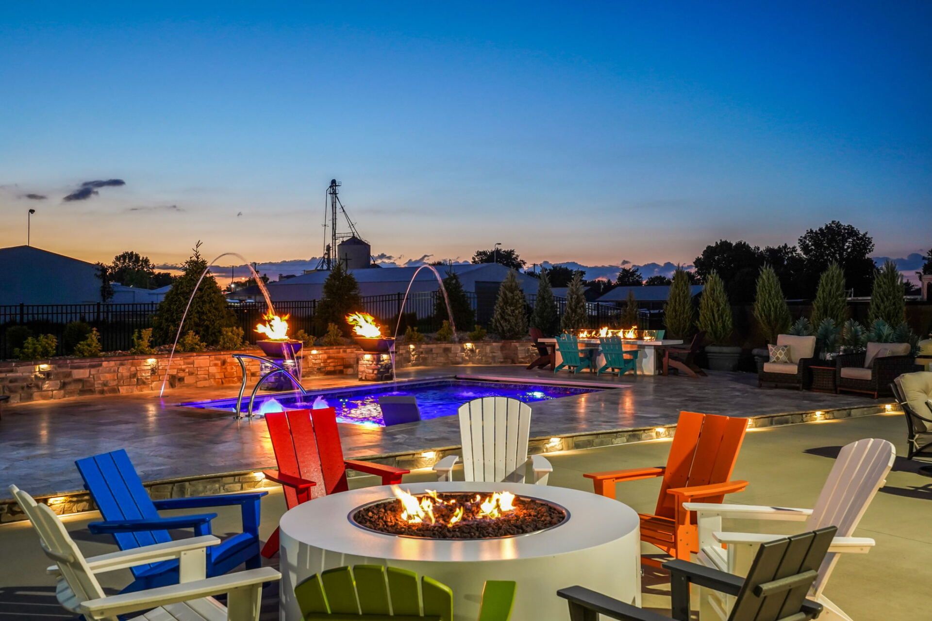 Colorful chairs around a fire pit beside a lit pool at sunset with lounge chairs and umbrellas