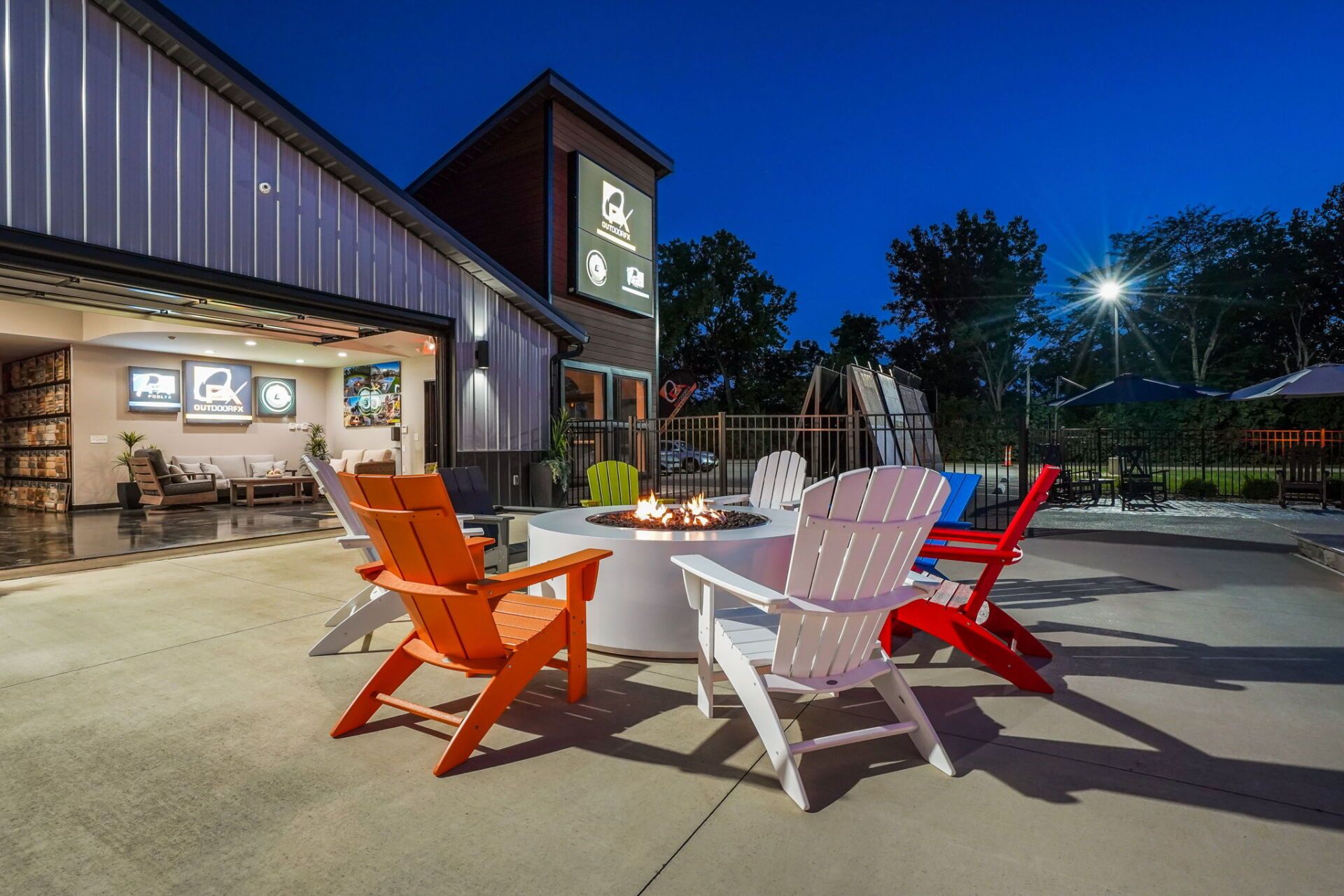 Outdoor patio with orange, white, and red chairs around a white table beside a lit building at dusk