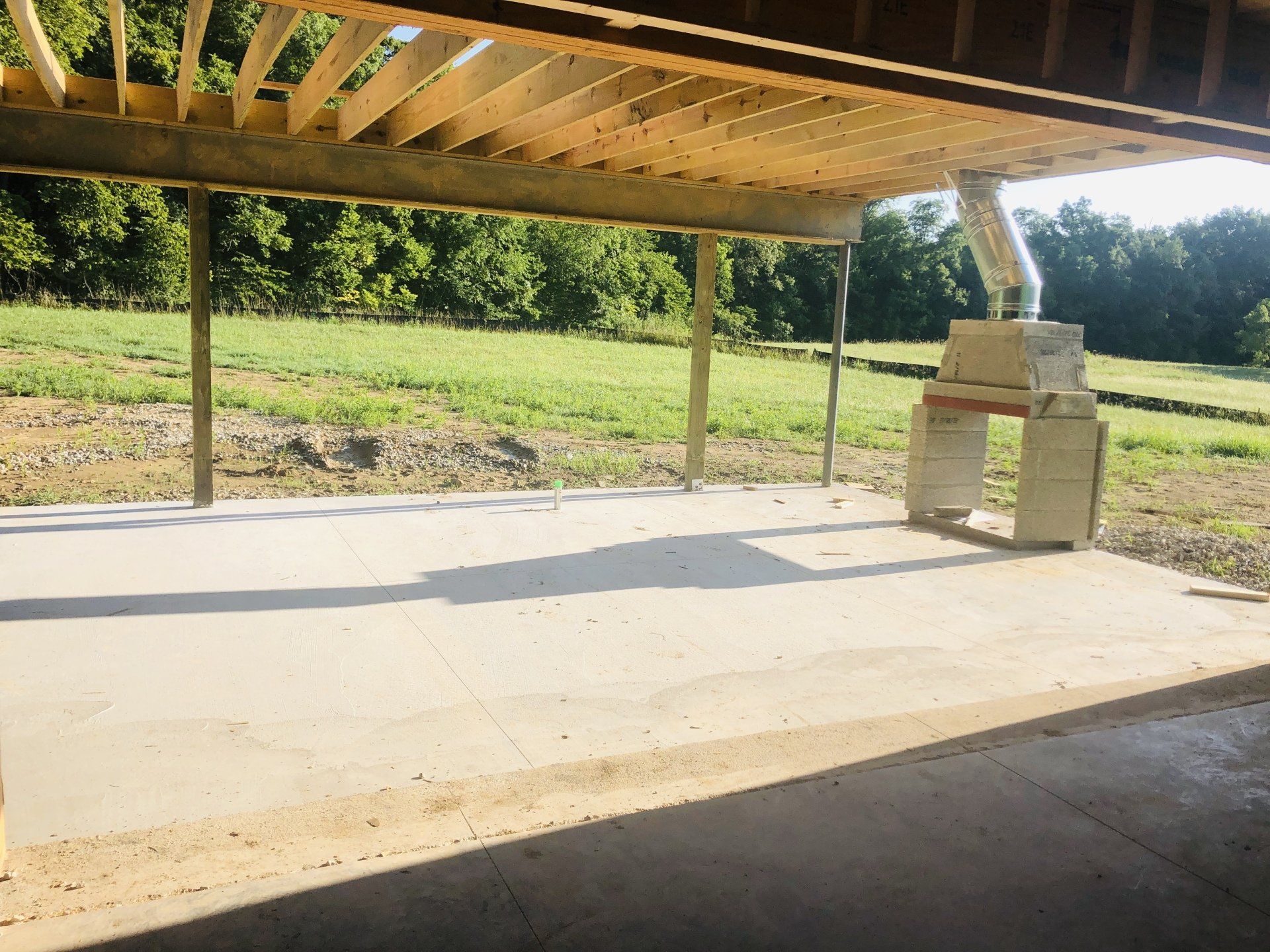 Wooden pergola over a concrete patio with a brick barbecue grill beside a grassy field.