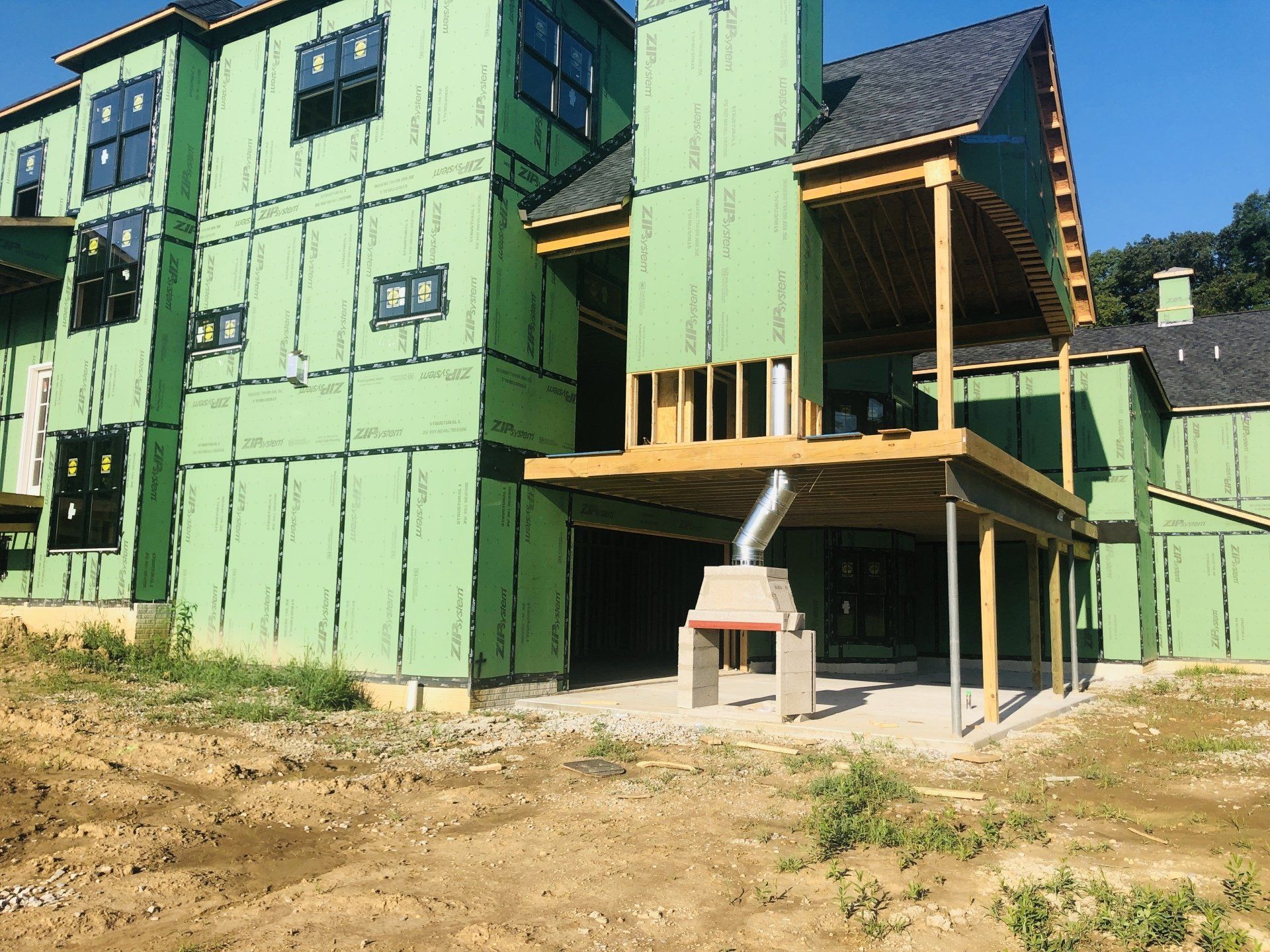 Partially built house with green exterior wrap, wooden porch framing, and bare dirt foreground