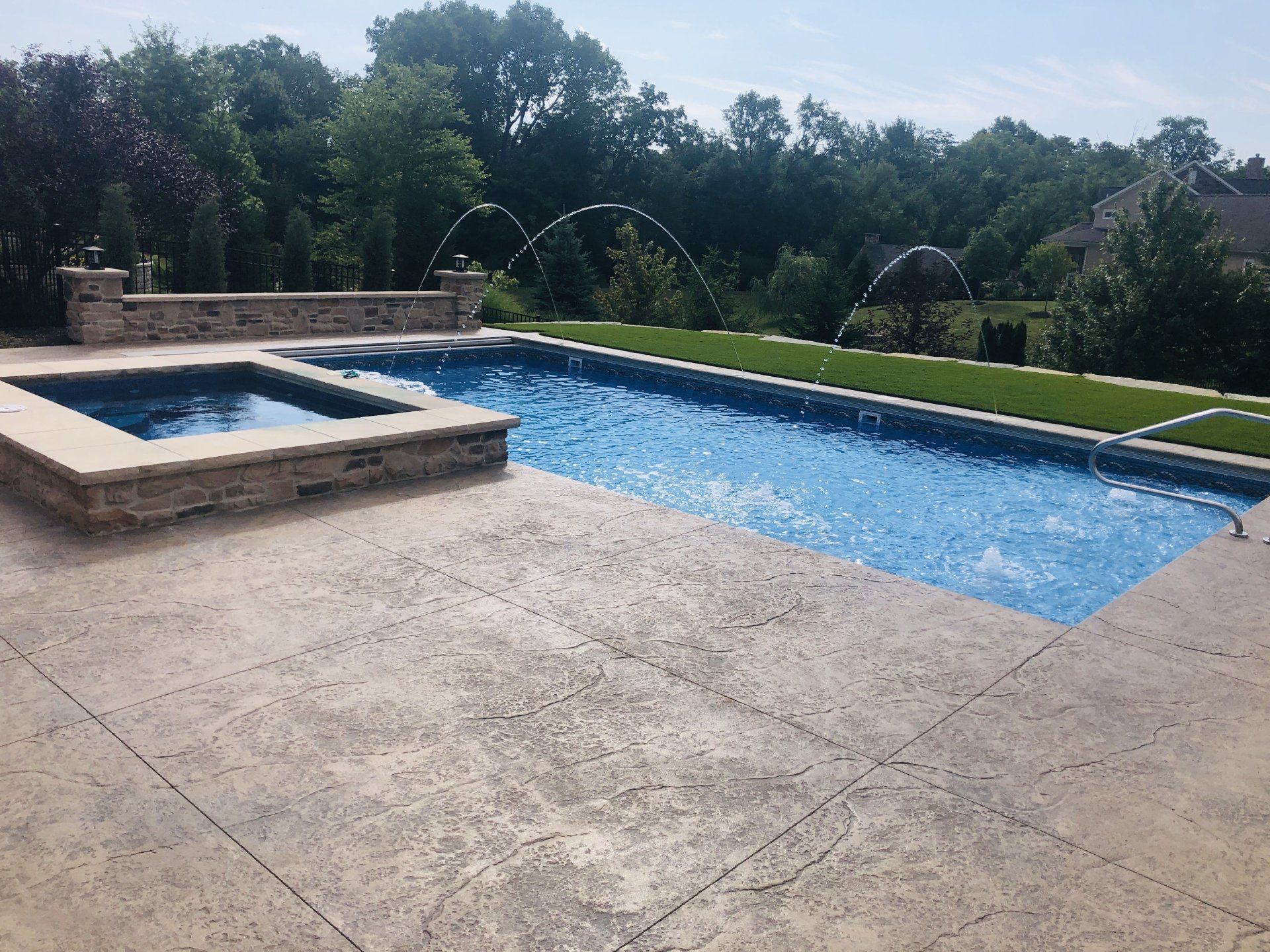 Backyard swimming pool with arcing water jets, stone hot tub, and manicured lawn under trees