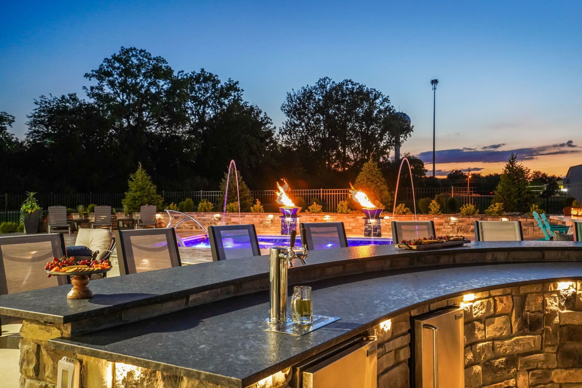 Backyard patio at dusk with stone fire pits, glowing lights, and trees under a blue-orange sky