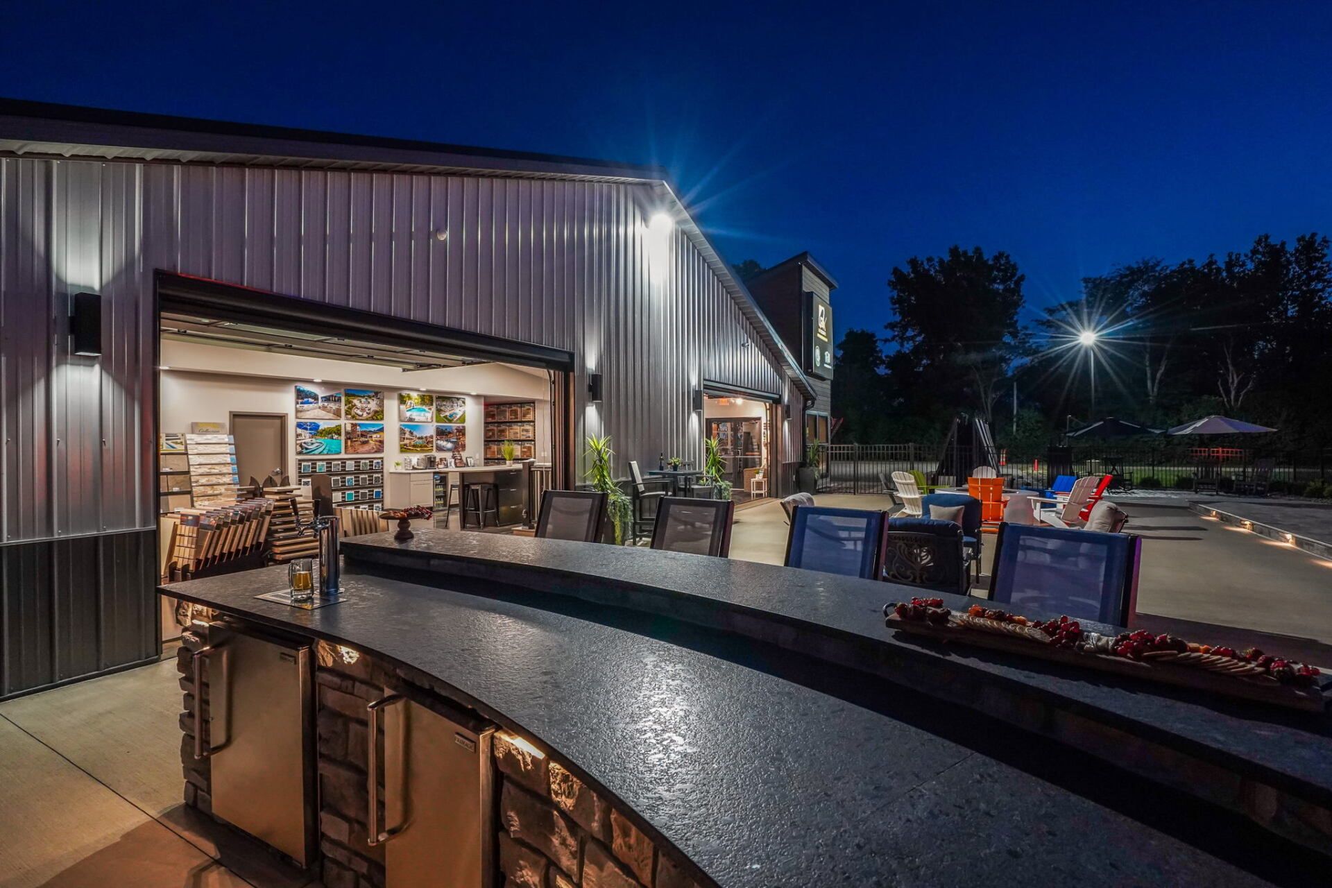 Outdoor bar and patio at dusk with metal building, glowing interior, and a curved counter under a deep blue sky