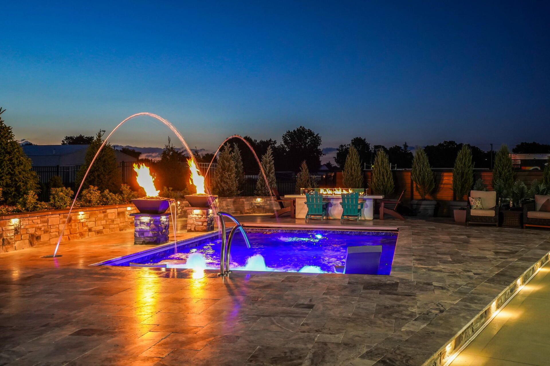 Outdoor patio with illuminated fountain and pool at dusk, surrounded by stone walls and lounge chairs