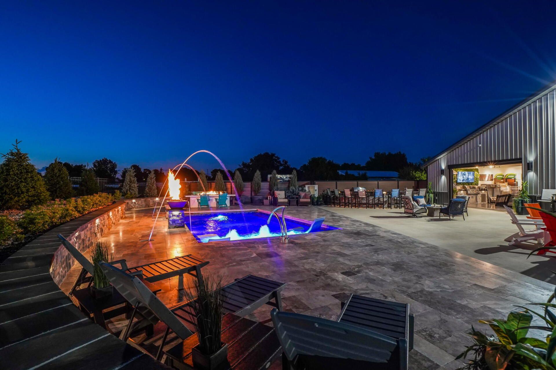 Nighttime patio with glowing blue pool, lounge chairs, and a building with warm lights
