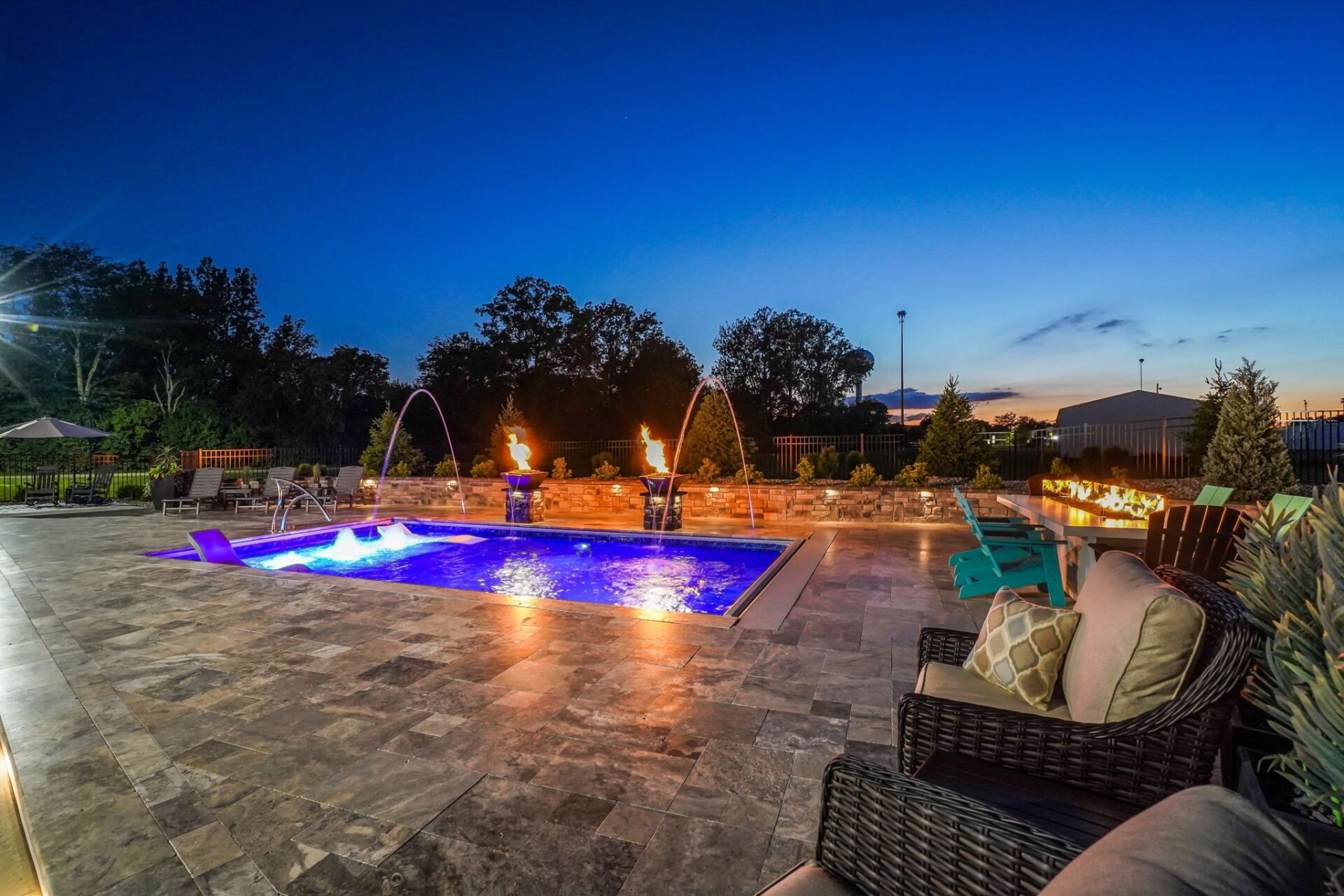 Patio at dusk with a glowing blue pool, wicker chairs, and warm garden lights under a twilight sky