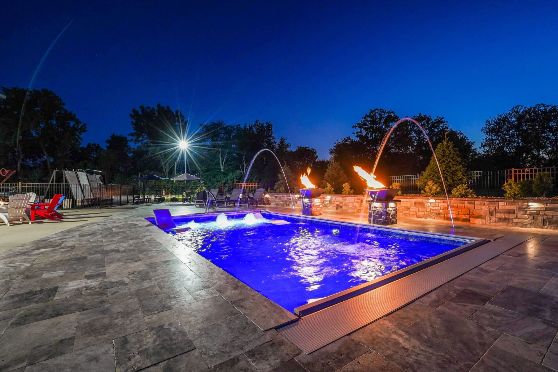 Illuminated backyard pool at night with blue water and fire features surrounding the deck