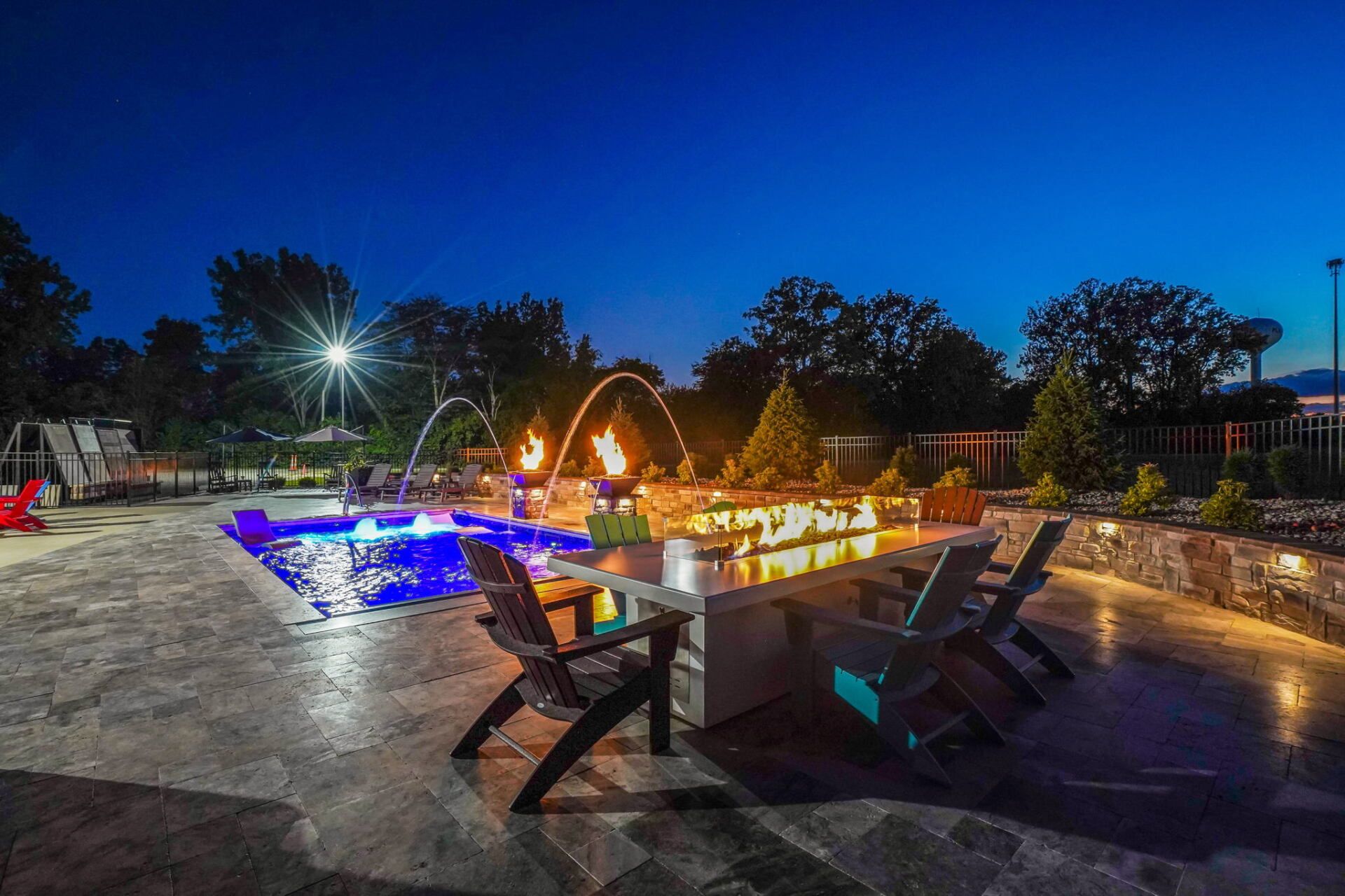 Outdoor patio at dusk with lit fire features, lounge chairs, and a blue-lit pool under a deep blue sky