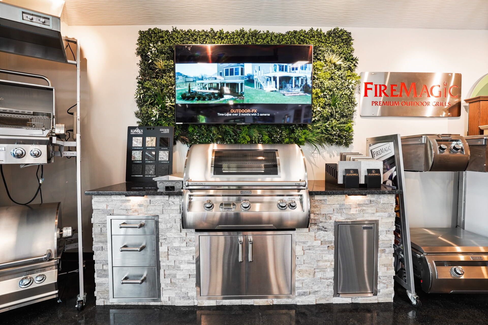Outdoor grill display with stainless steel barbecue, TV, and green hedge backdrop inside a showroom