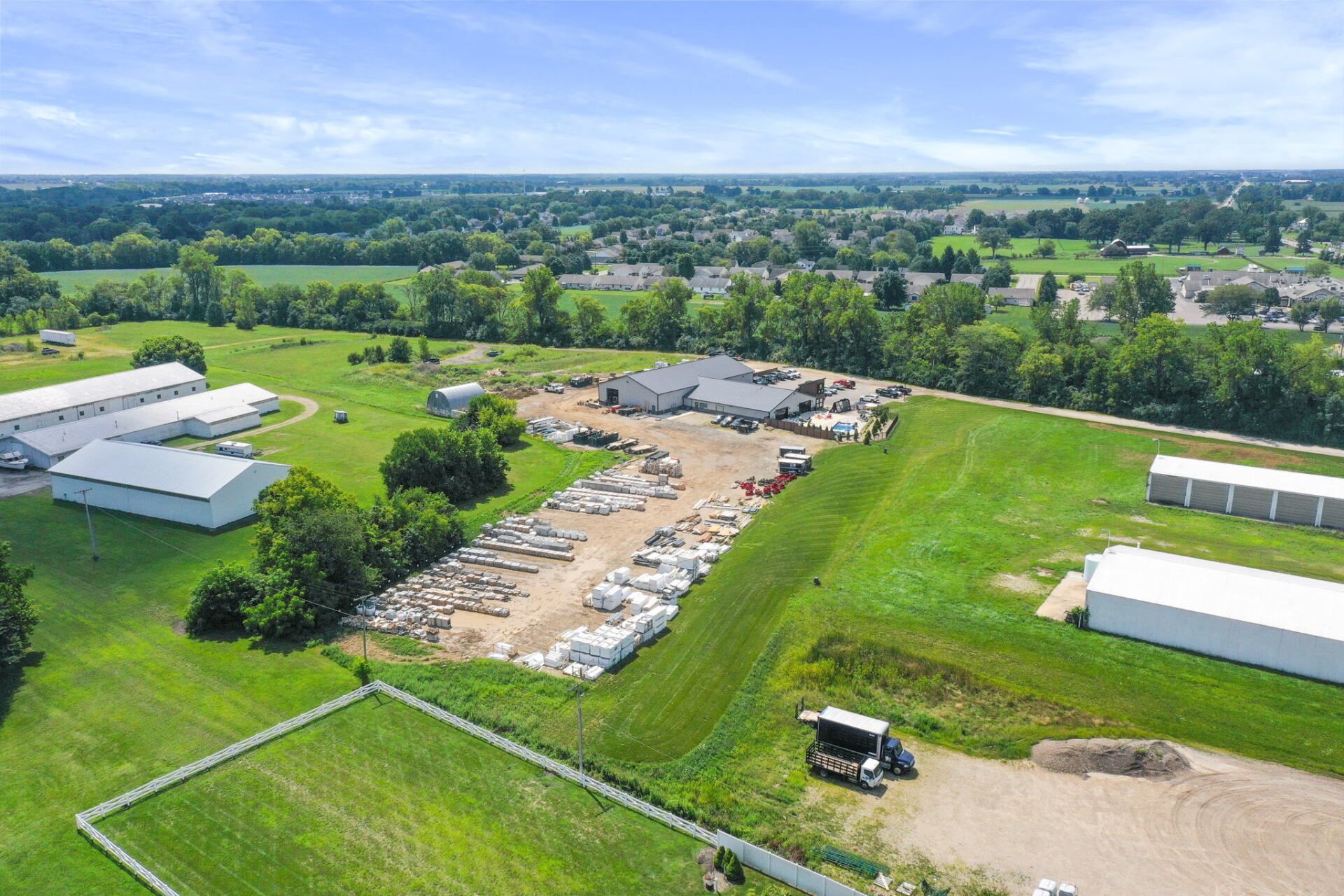 Aerial view of a rural farmyard with barns, parked vehicles, and green fields under a clear sky