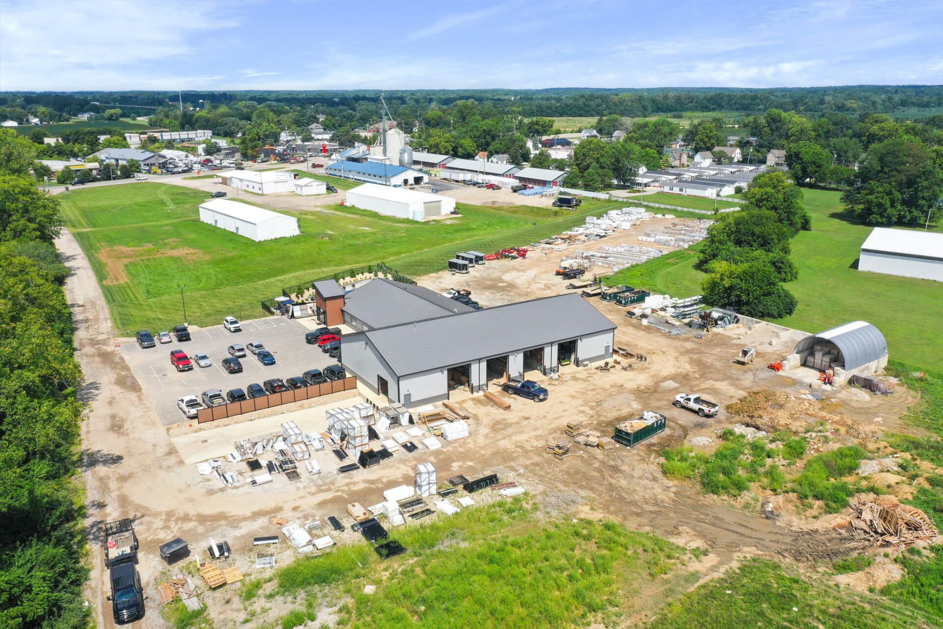Aerial view of a construction site with a gray-roofed building, equipment, dirt piles, and surrounding green fields.