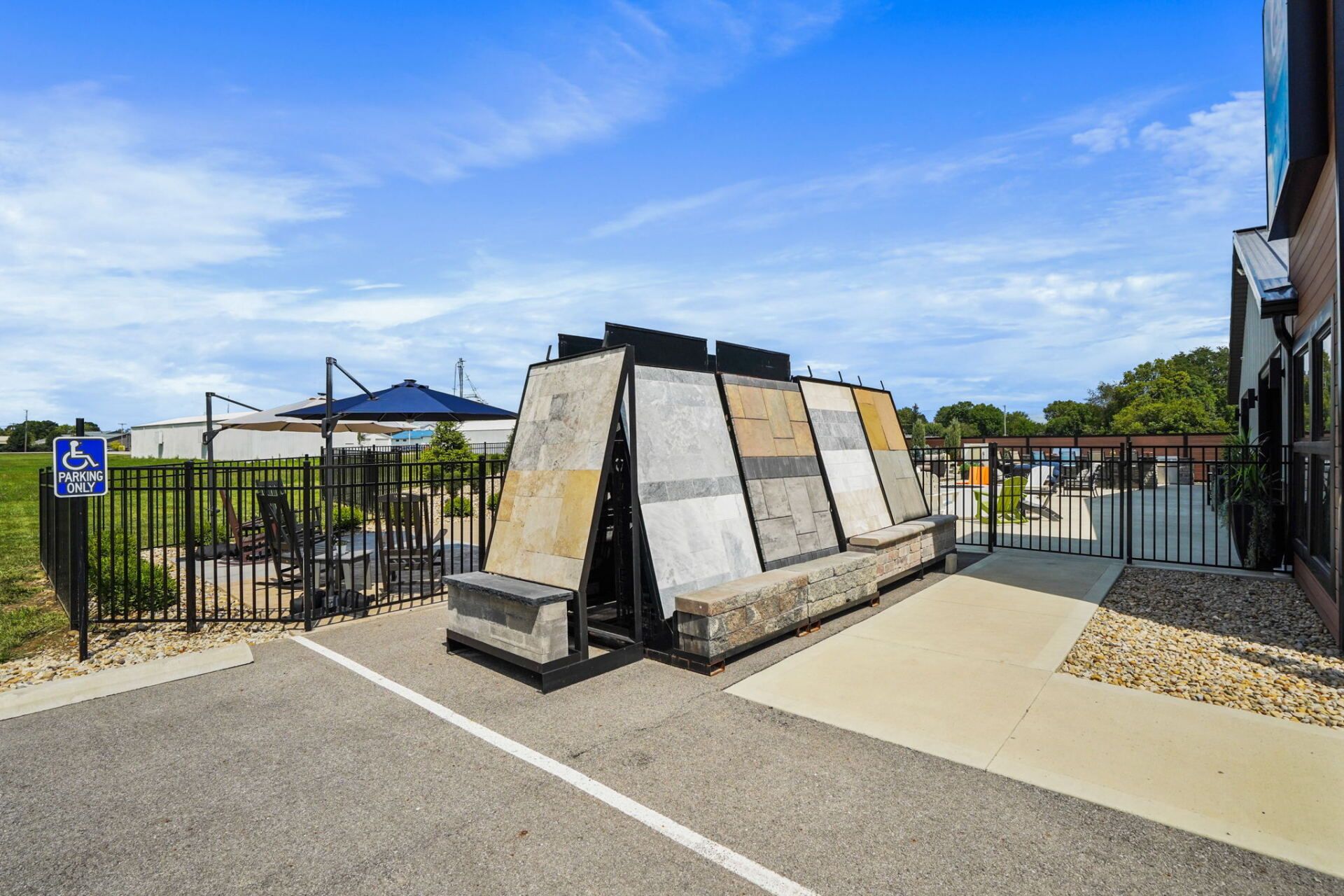 Outdoor fenced display of large tilted stone panels beside a paved walkway under a blue sky