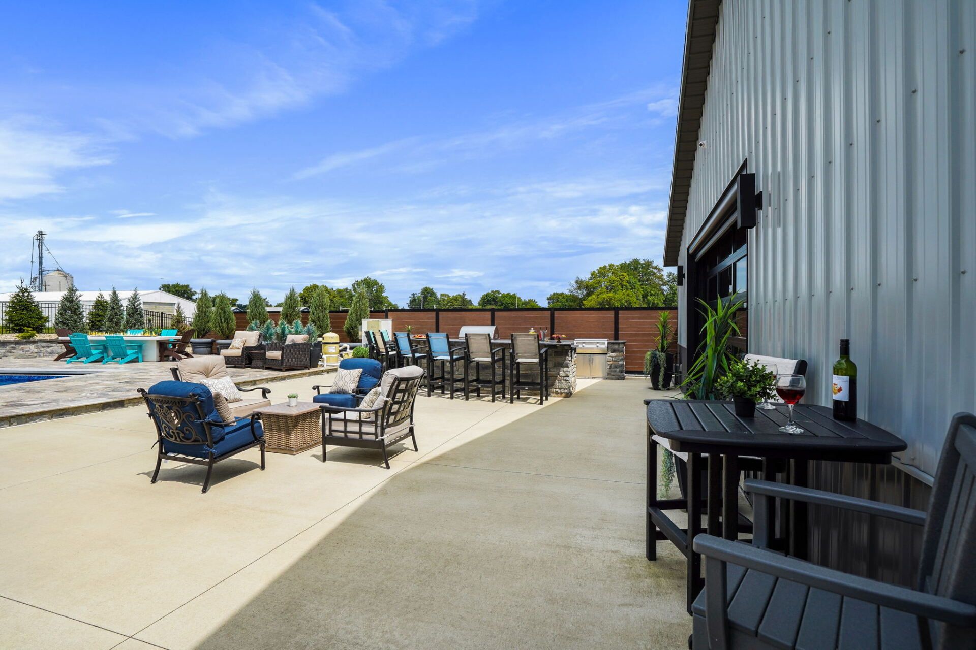 Outdoor patio with tables and chairs beside a pool, next to a corrugated metal building under a blue sky
