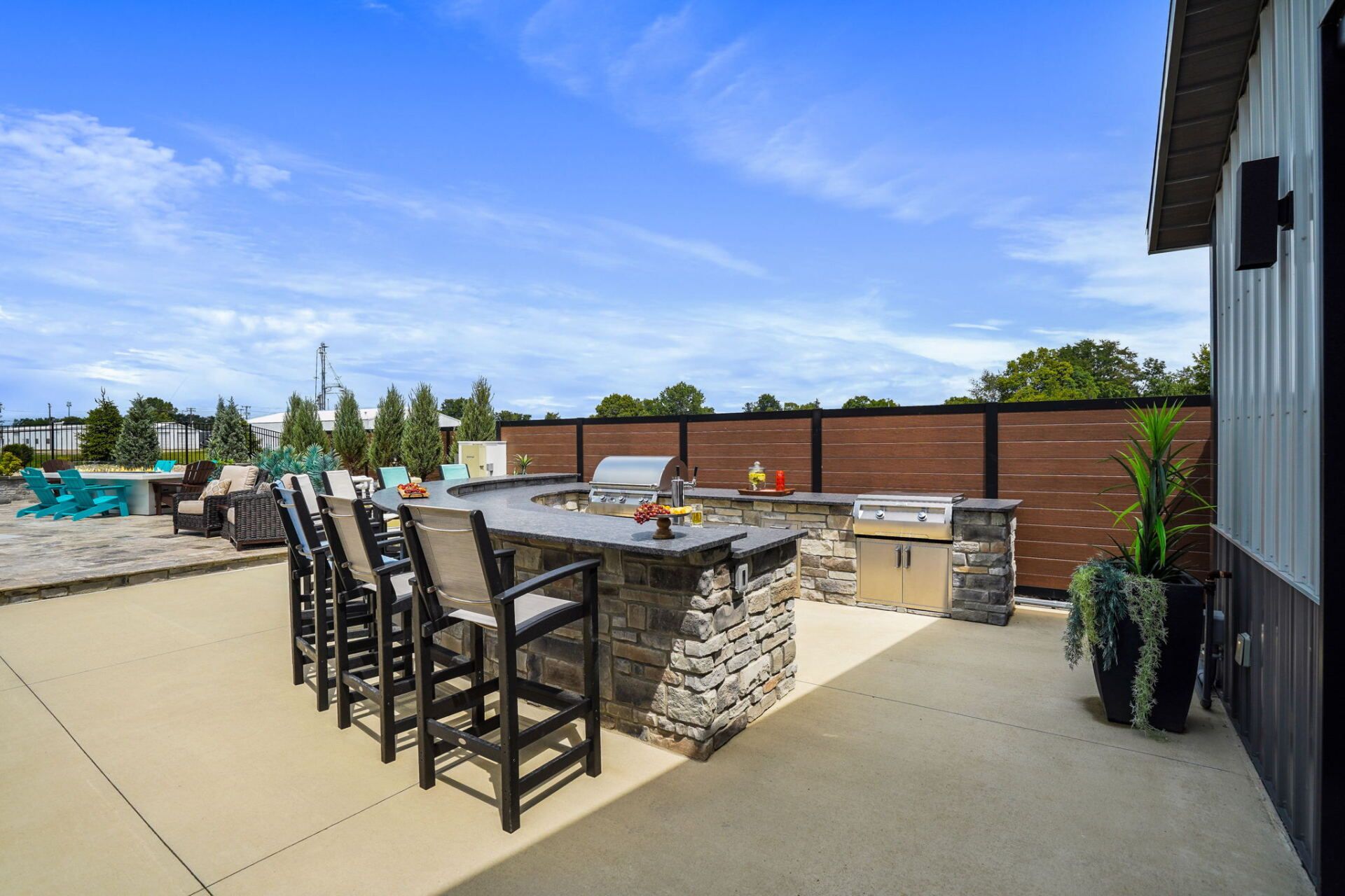 Outdoor patio with stone bar, tall stools, grill, and potted plants under a bright blue sky.