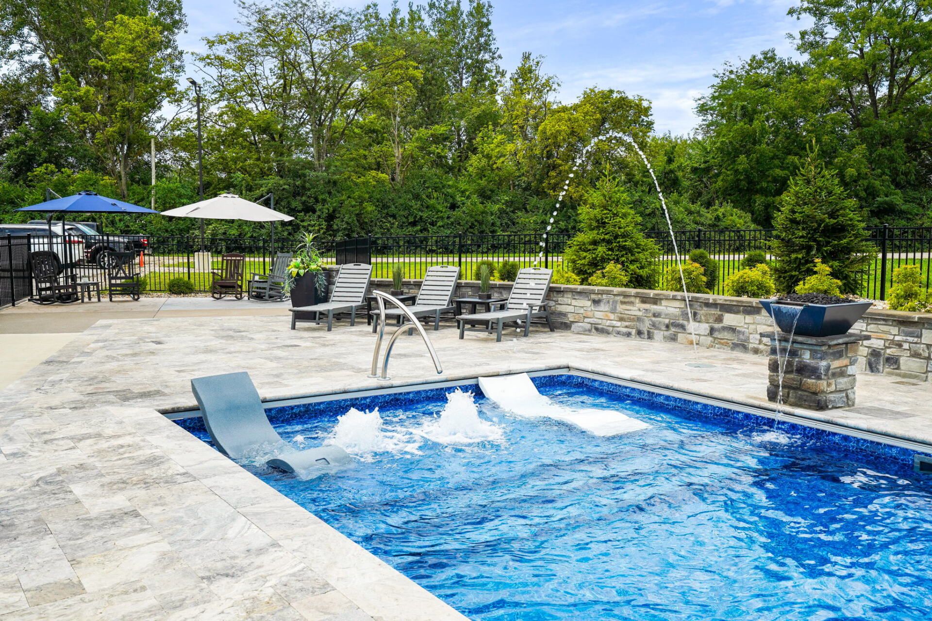 Backyard pool with blue water, stone deck, lounge chairs, umbrellas, and lush green trees